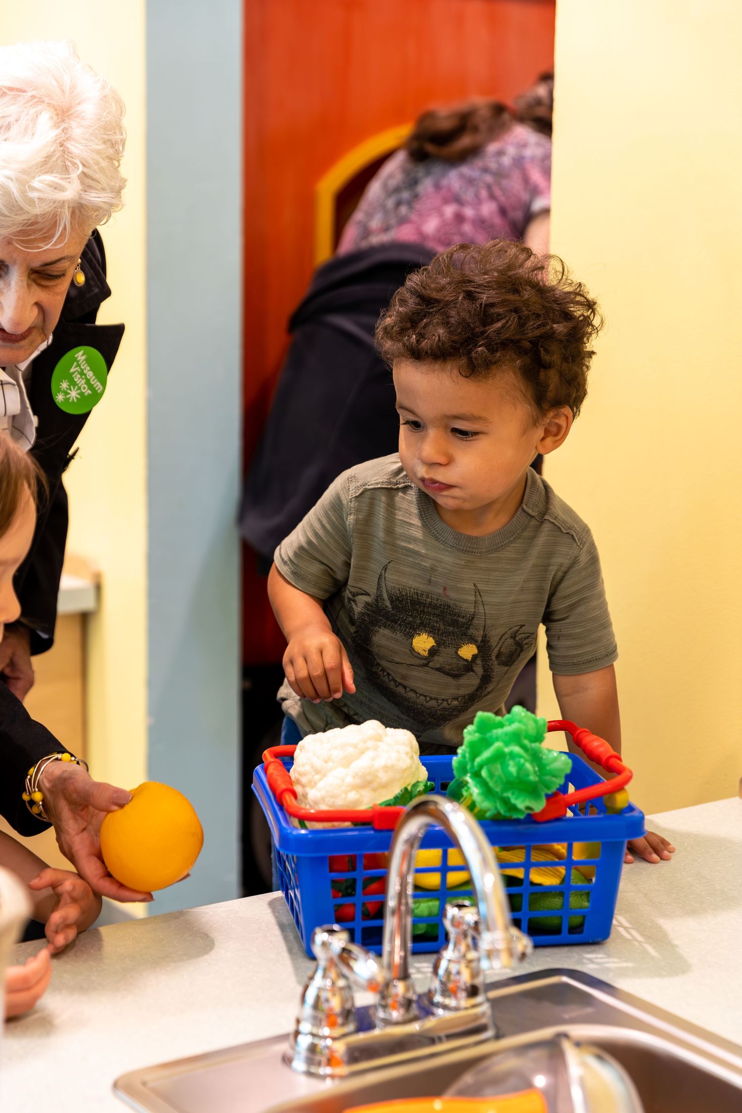 Child playing with colorful toys in basket by sink as adult looks on at educational or daycare setting.