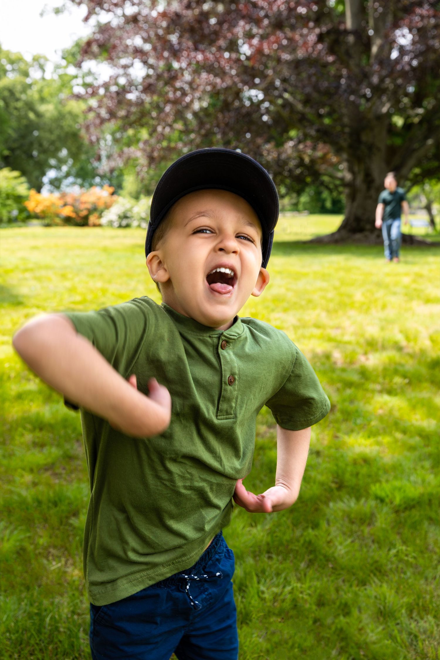 Child in green polo shirt and baseball cap plays energetically on a sunny day in a grassy backyard with flowers.