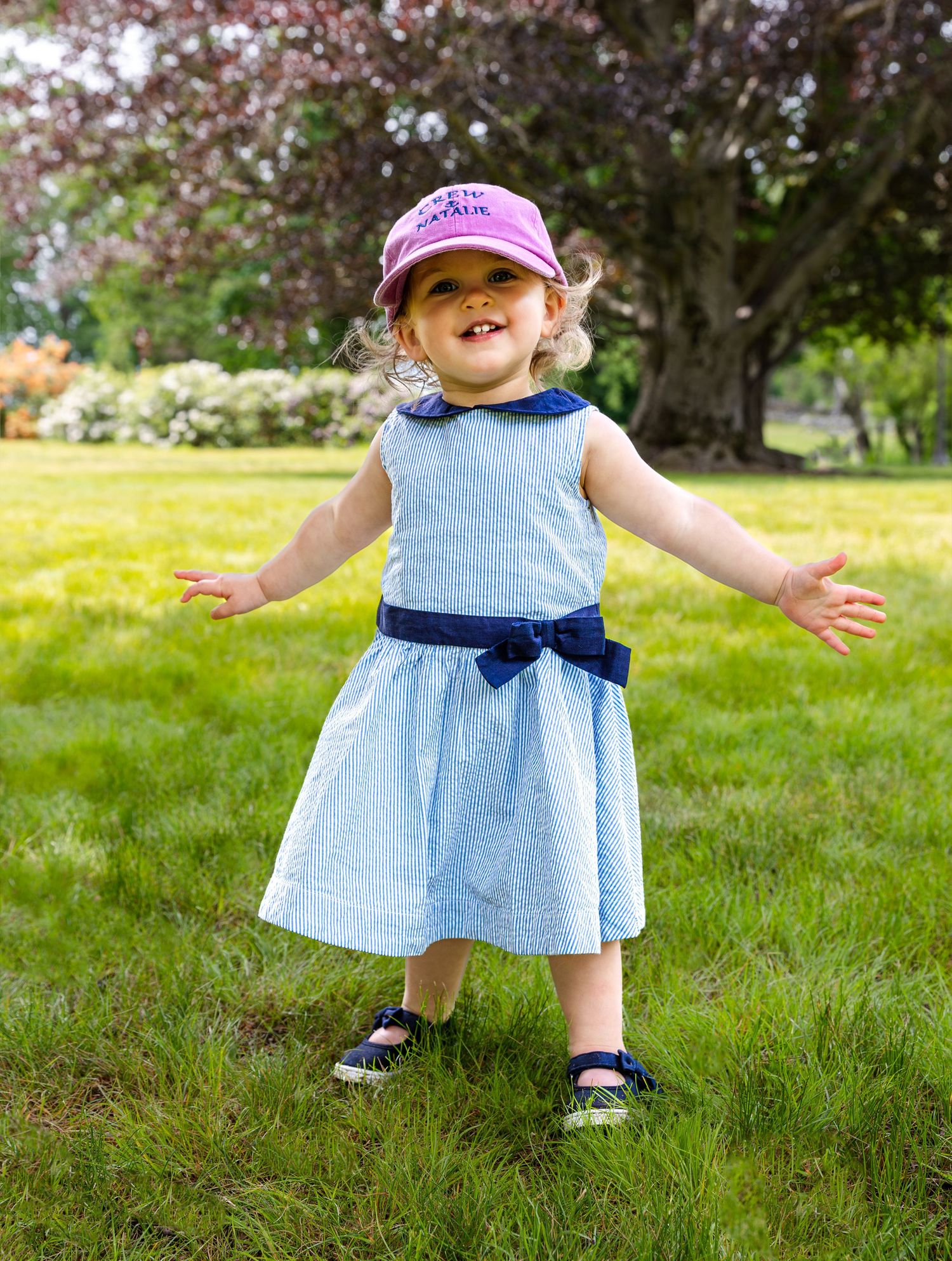 A toddler in a light blue dress with navy bow explores a spring garden setting with blooming trees in background.