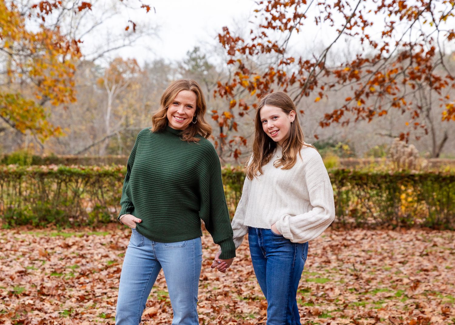 Two people wearing sweaters stand together outdoors on fallen autumn leaves surrounded by orange fall foliage.