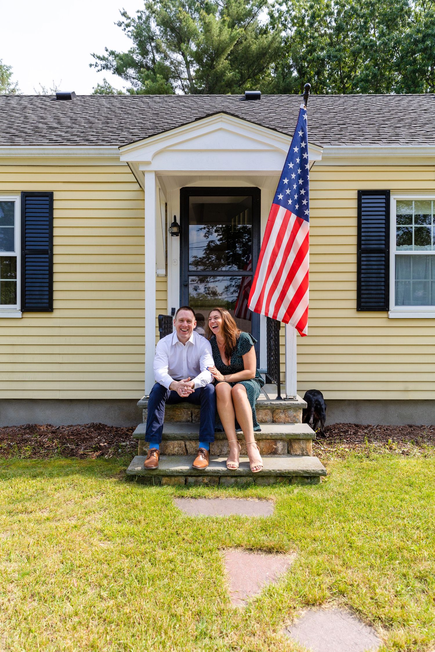 A couple sits on the front steps of a yellow house with black shutters and an American flag displayed by the door.
