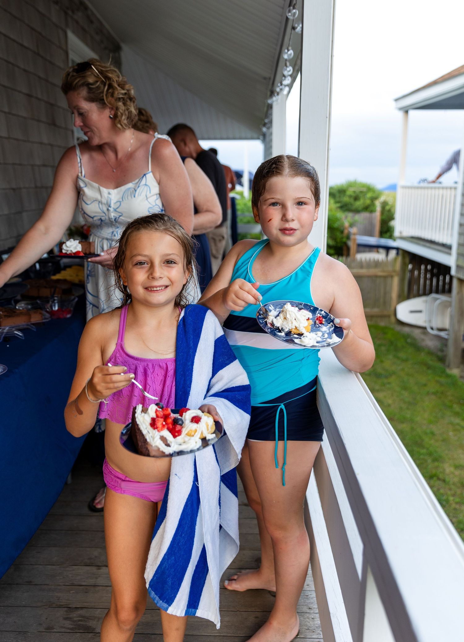 Two kids in swimsuits holding dessert plates with ice cream on a beachfront porch during summer.