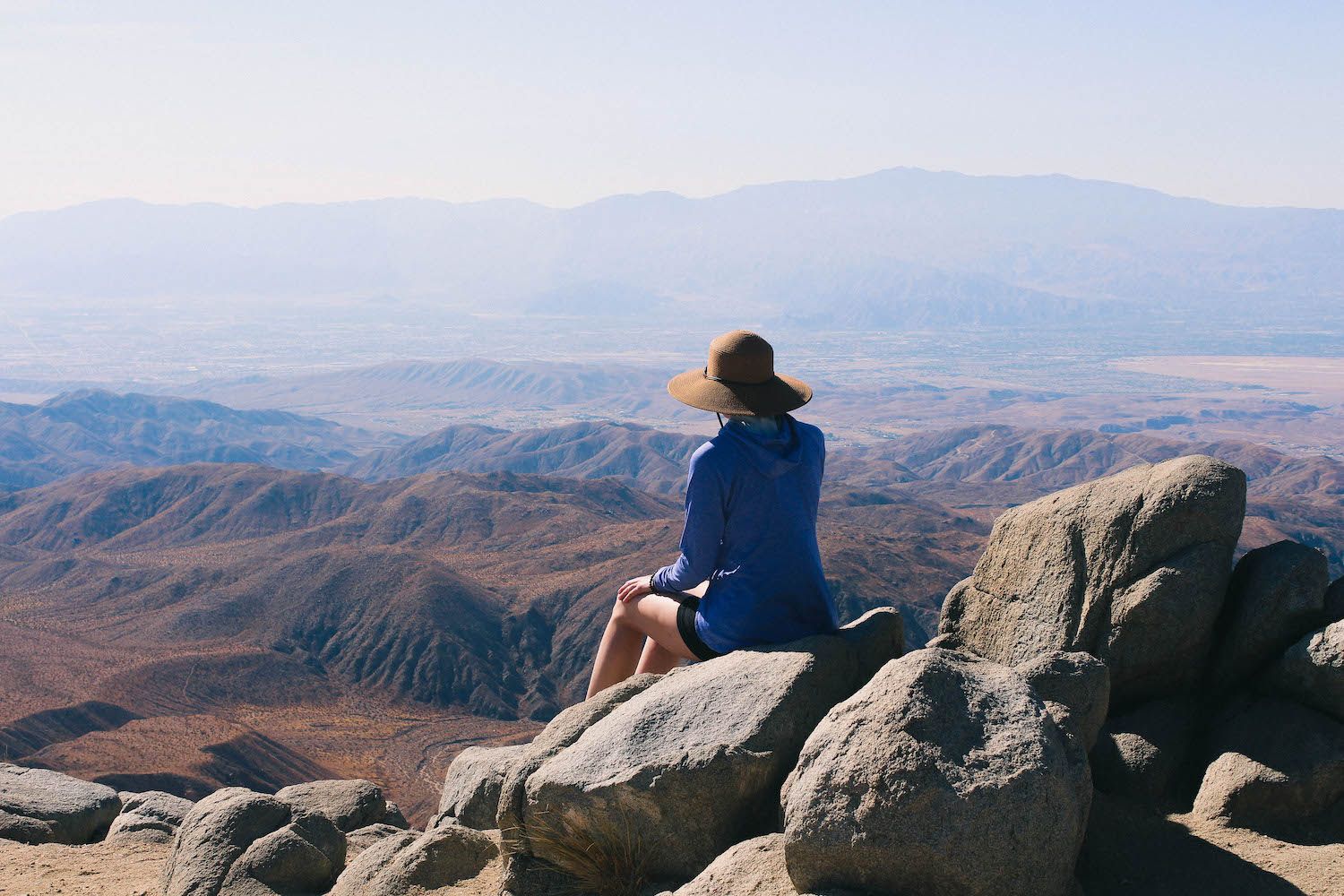 A hiker sits on rocky outcrop overlooking vast mountain ranges and valleys in the distance.