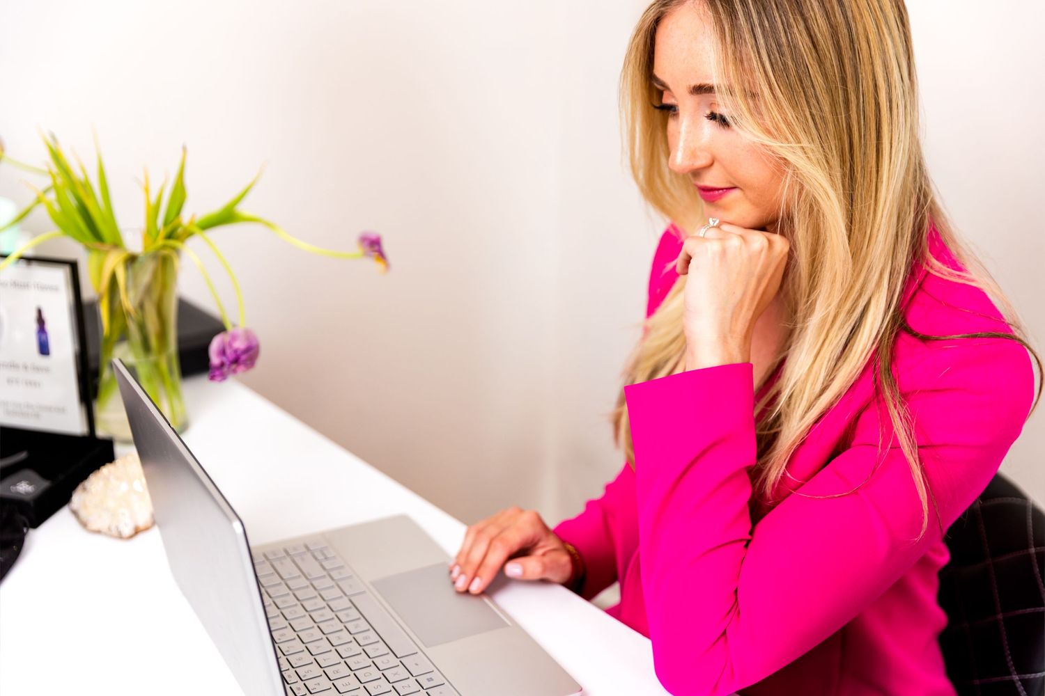 Person in bright pink blazer smiling while working on laptop in home office setting.