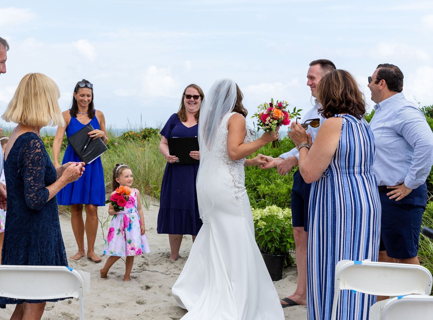 Wedding guests gathered on a beach in blue and white attire watching a celebration by the ocean.