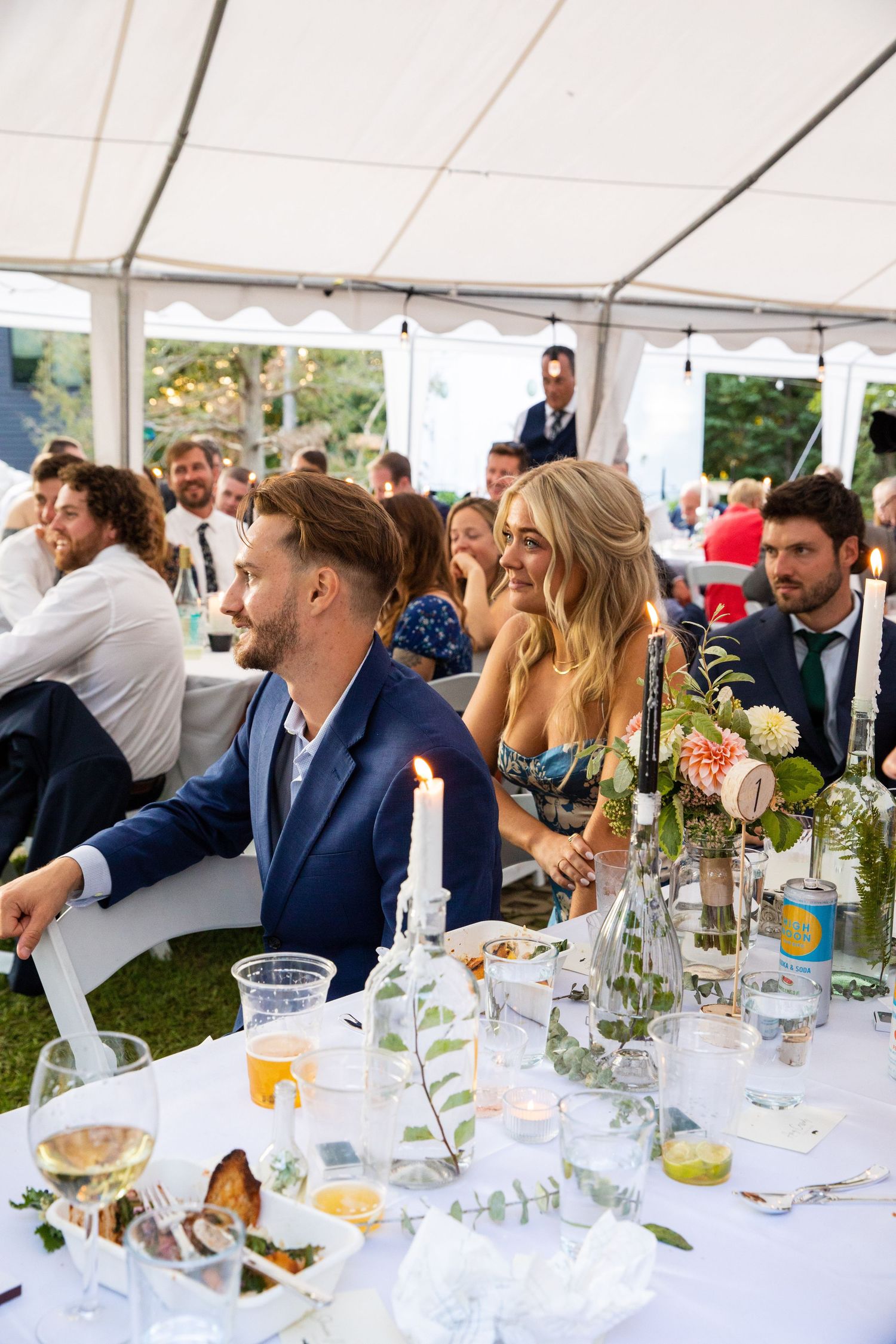 Wedding guests enjoy a reception dinner under a white tent with elegant table settings and floral centerpieces.