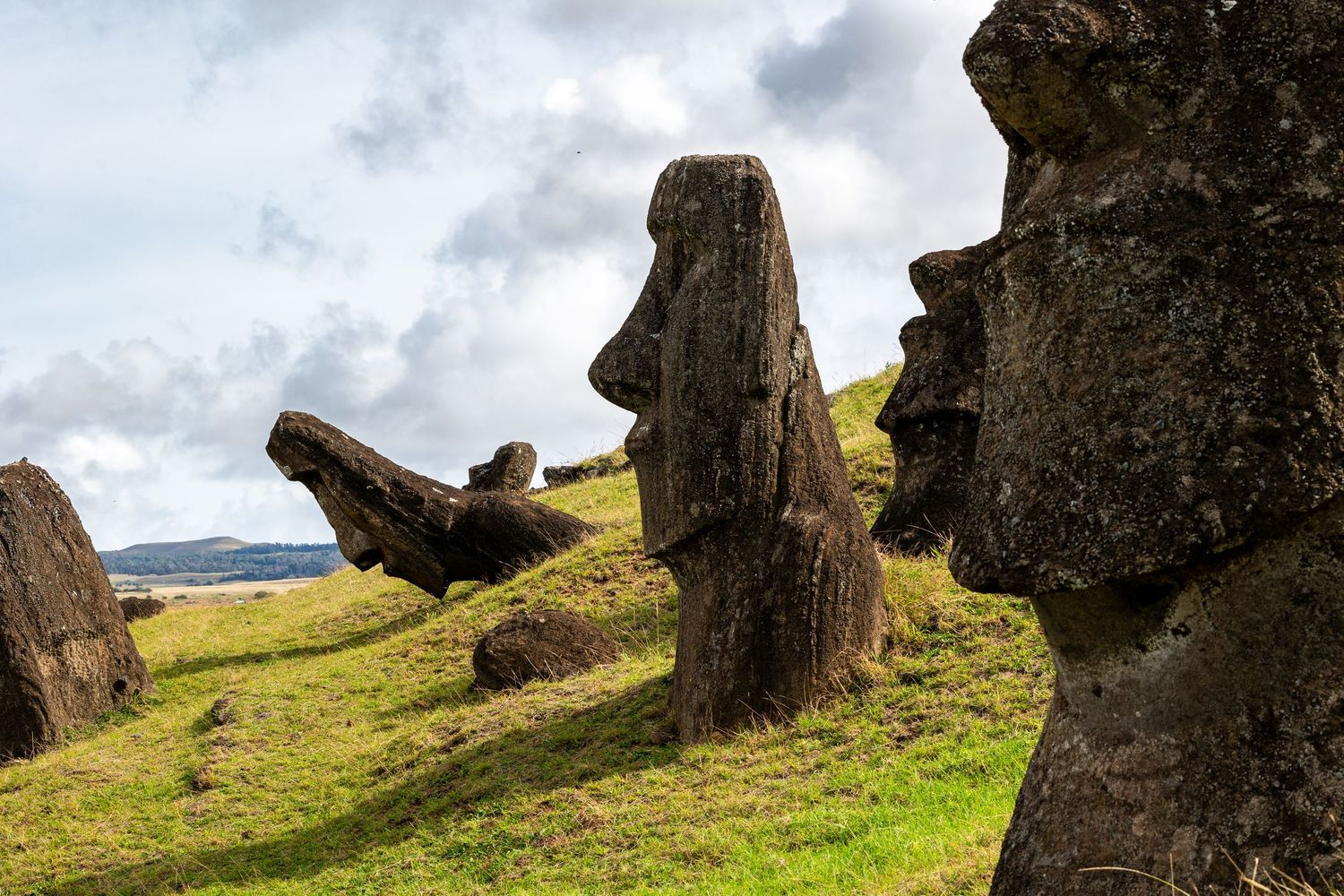 Ancient moai stone statues stand scattered on the green hills of Easter Island under a cloudy sky.