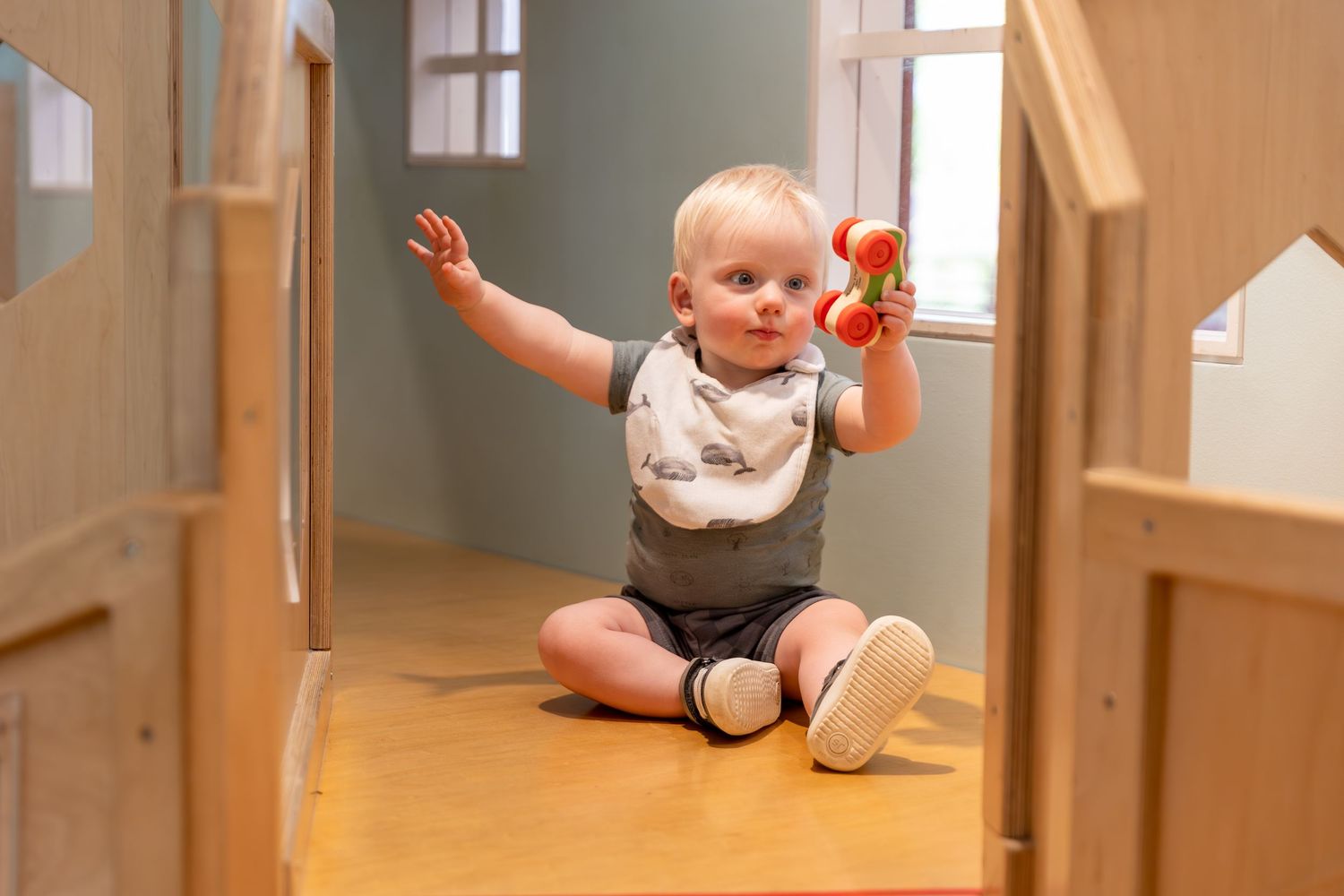 A toddler crawls across a sunlit hardwood floor in a bright hallway with white walls and windows in view.
