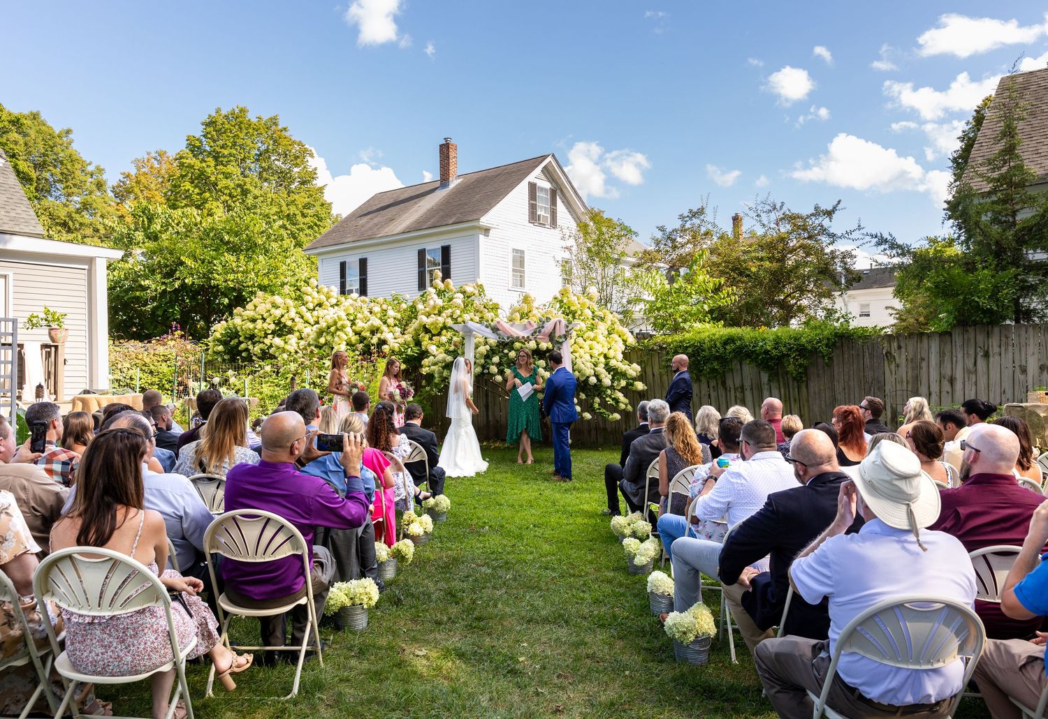 Panoramic view of guests sitting in white chairs at an outdoor garden wedding ceremony on a sunny day.