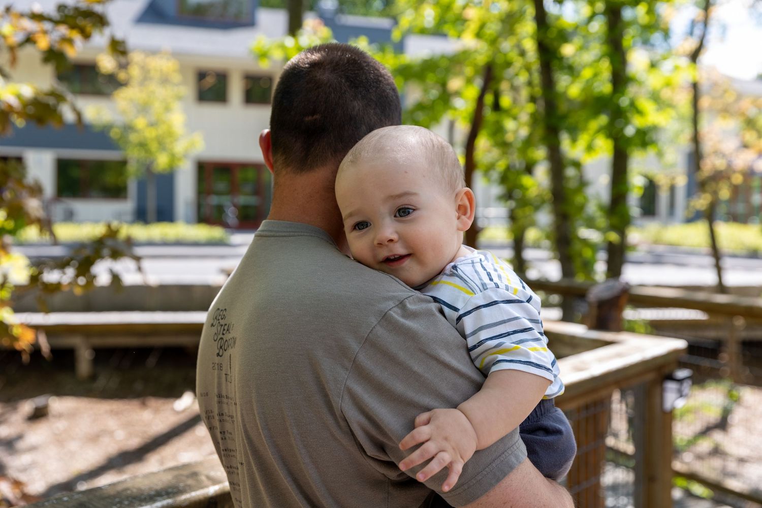 Baby in striped shirt peeks over shoulder while being held outdoors on sunny day with picnic tables in background.