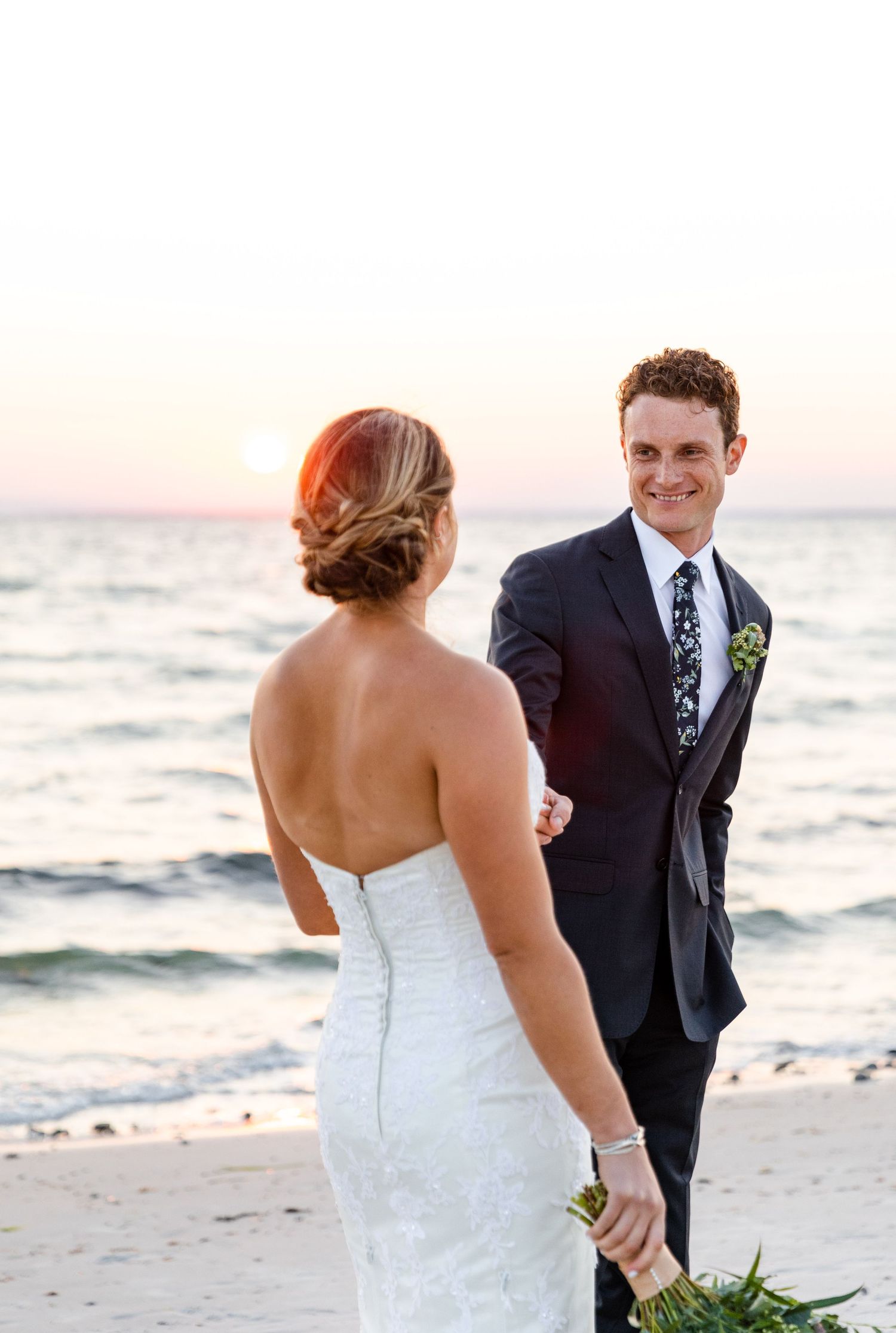 A couple shares a romantic moment on a beach at sunset, with the bride in a strapless white gown and groom in navy suit.
