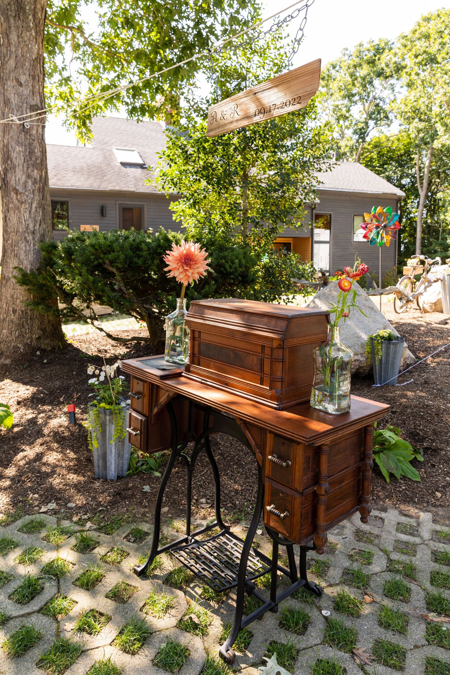 Vintage wooden piano with potted flowers sits on a patterned stone patio surrounded by lush greenery and dappled sunlight.