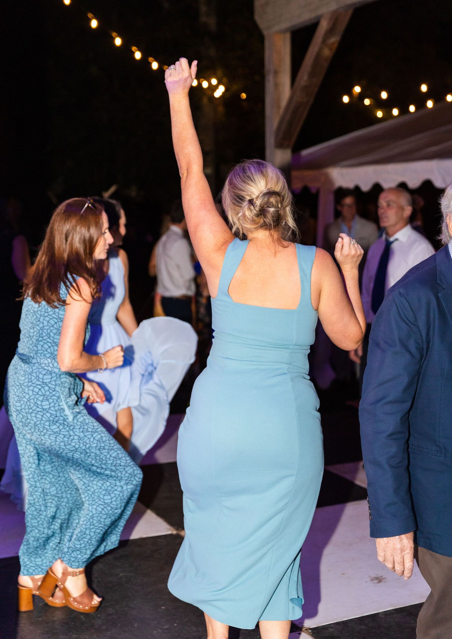 Two women in blue dresses dancing together at an outdoor evening reception with string lights overhead.