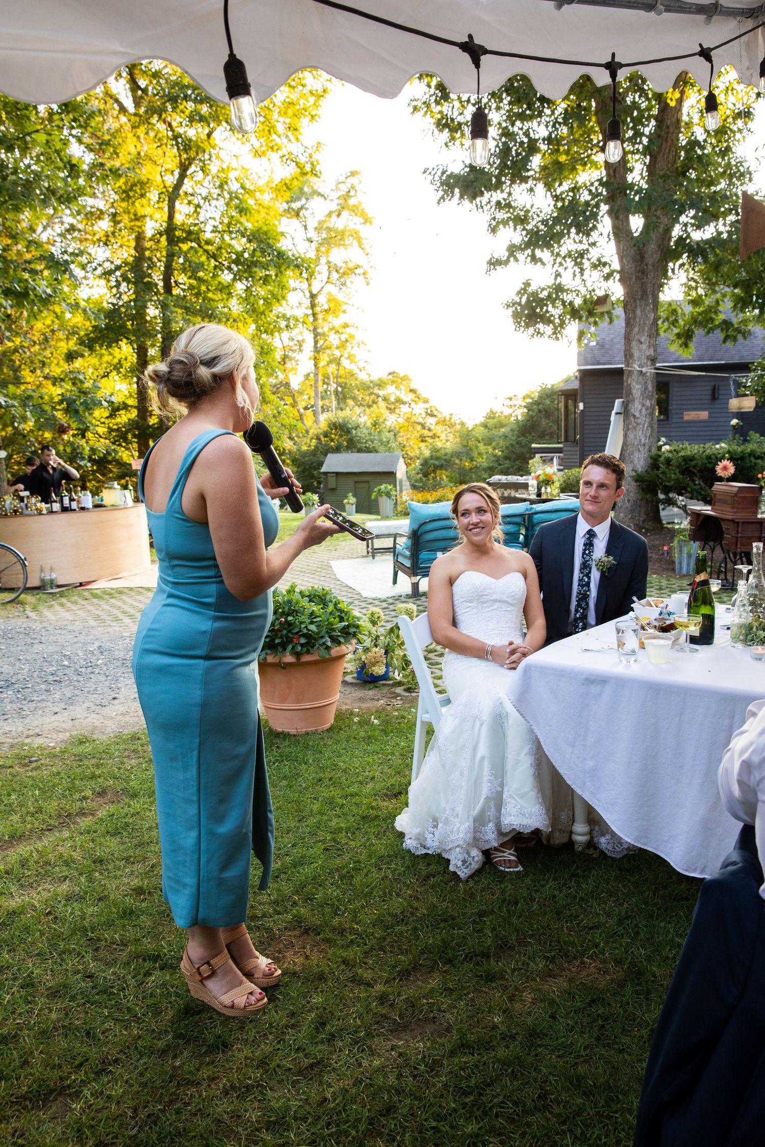 A person in a teal dress gives a wedding speech outdoors while the couple listens at an elegantly decorated table.