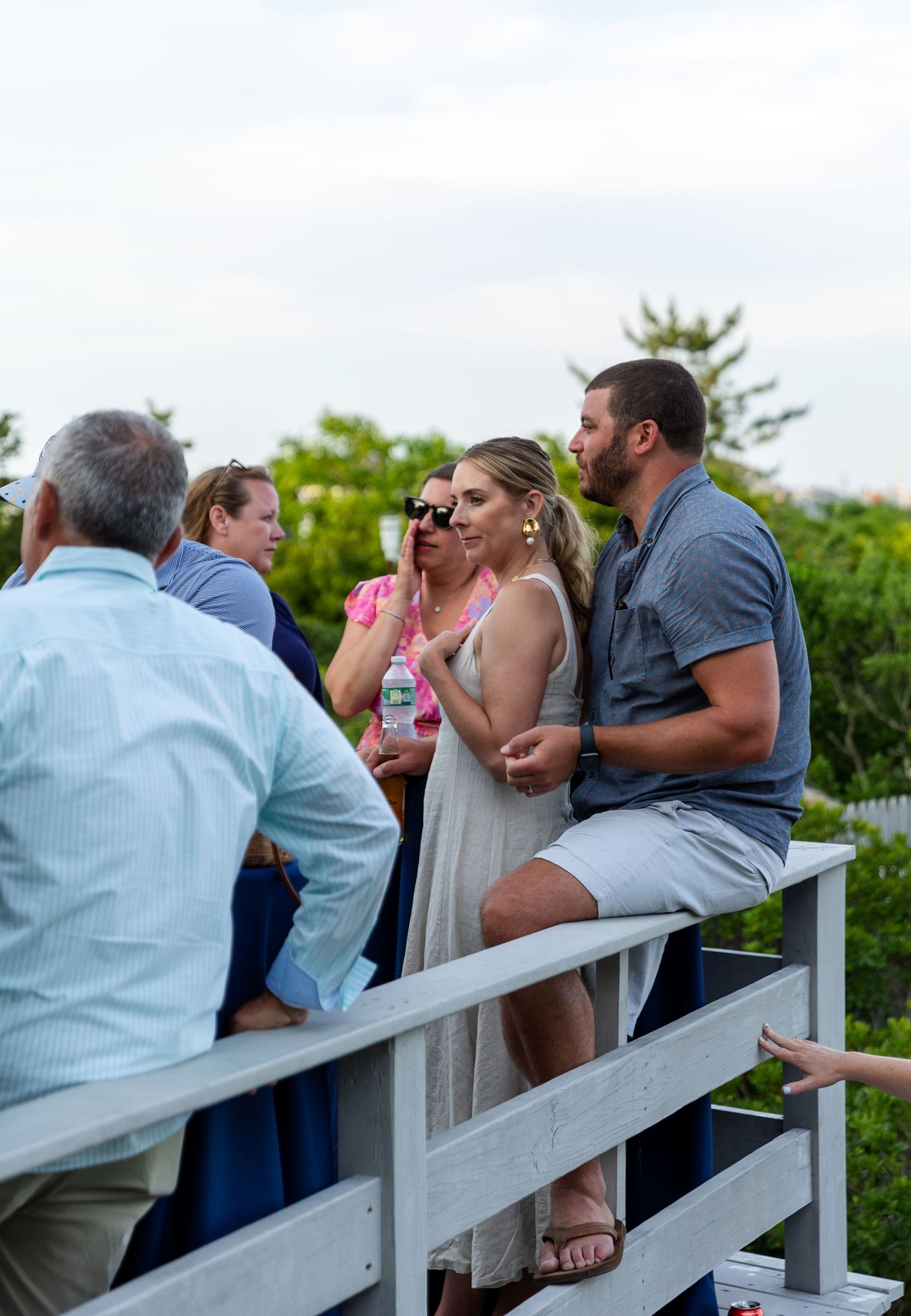 A group of people gather and socialize on a white railing deck outdoors during a casual summer gathering.