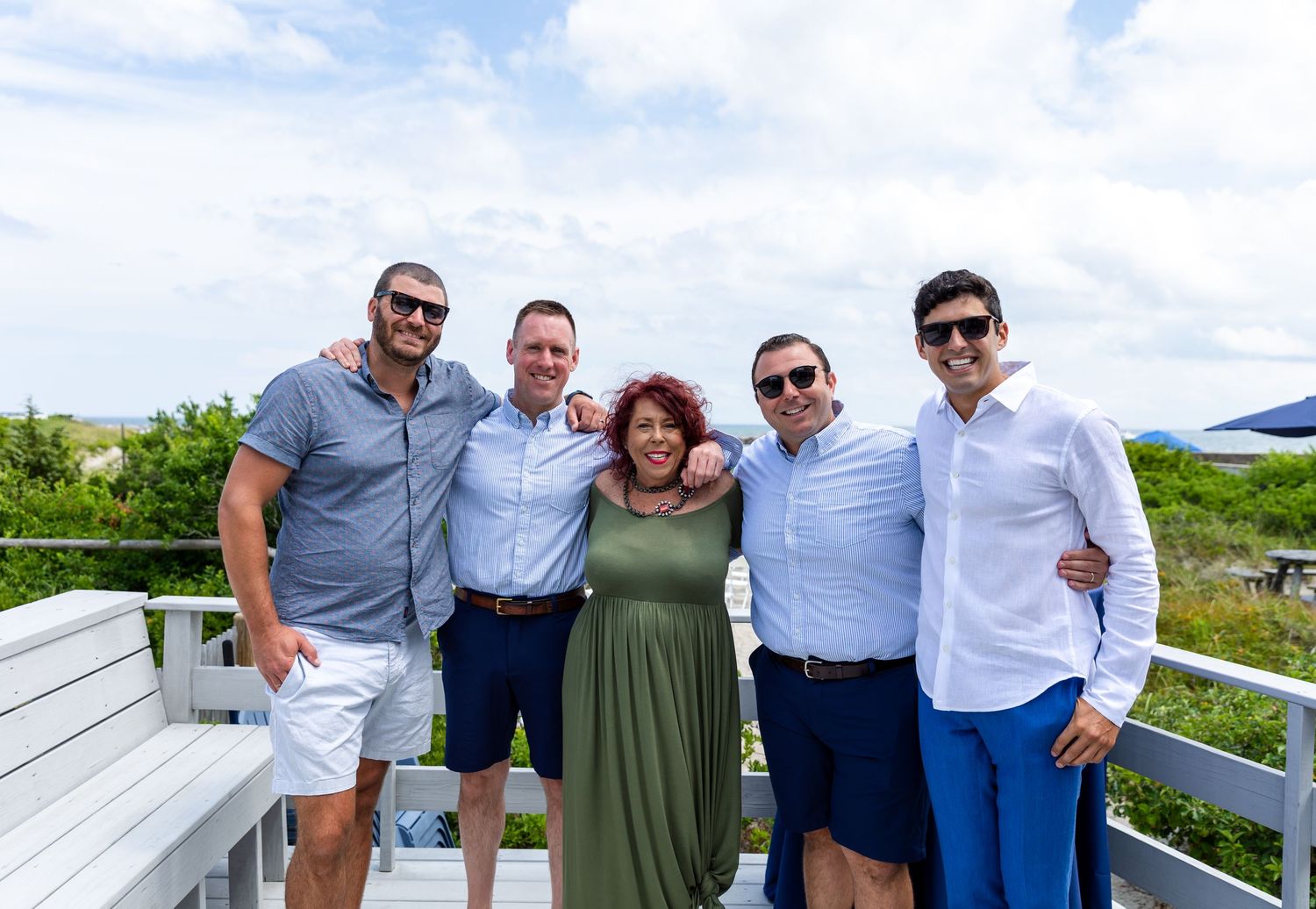 Group of people gathering outdoors on a wooden deck against a cloudy sky with bright sunlight.