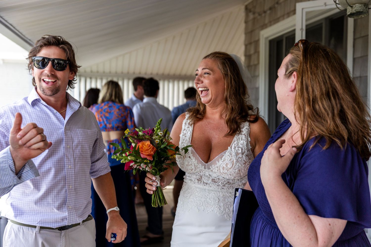 Candid wedding party moment on outdoor porch with guests in white and navy blue laughing together during celebration.