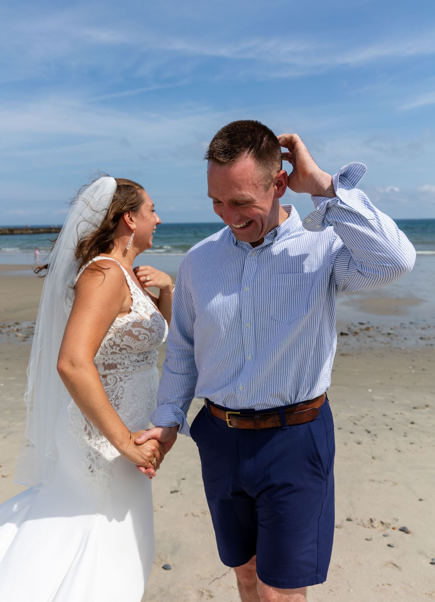 Happy couple sharing a candid moment on a sunny beach, with the bride in a white lace gown and veil.