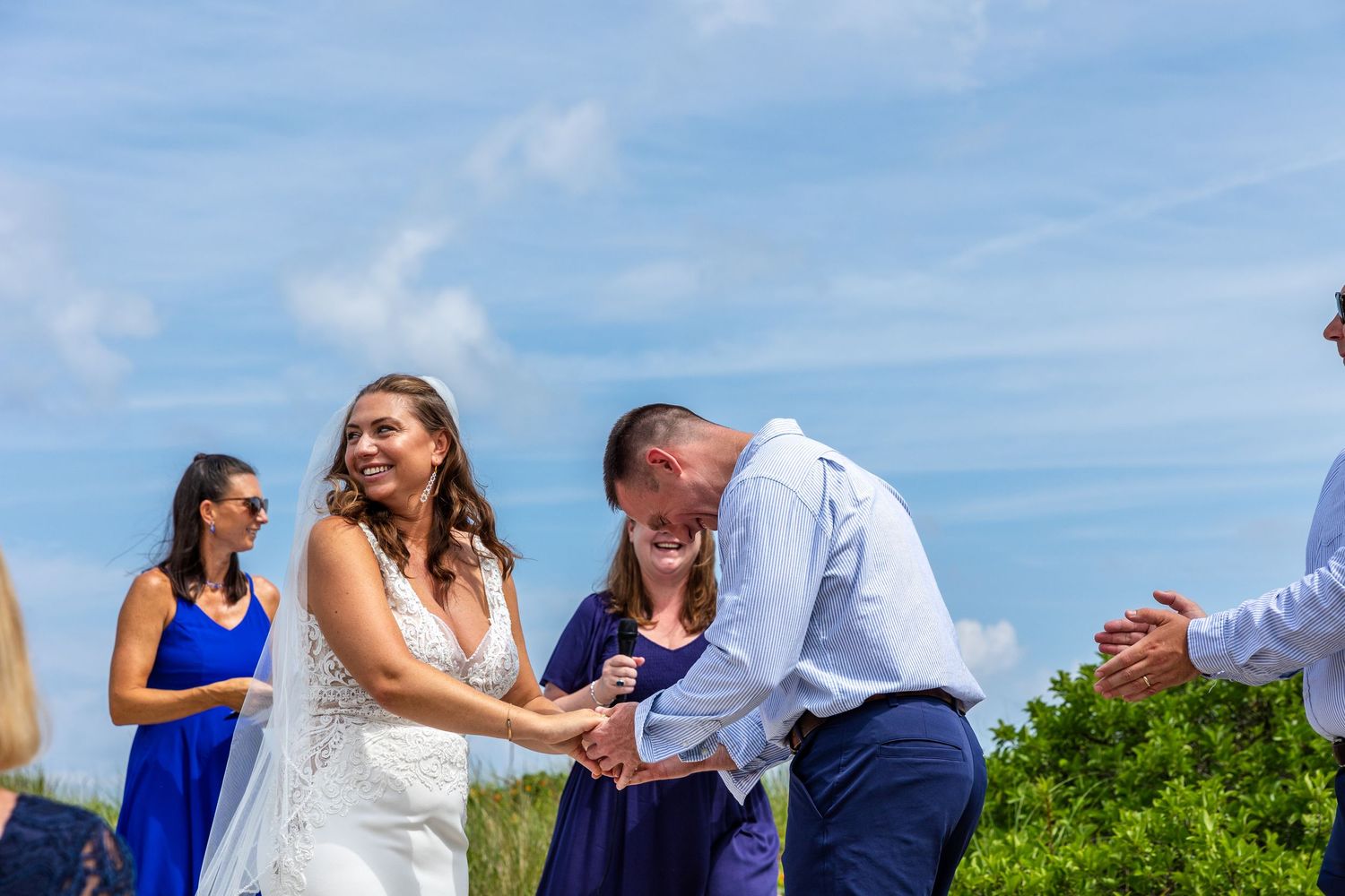 Wedding ceremony taking place outdoors on a sunny day with blue sky and green foliage in the background.