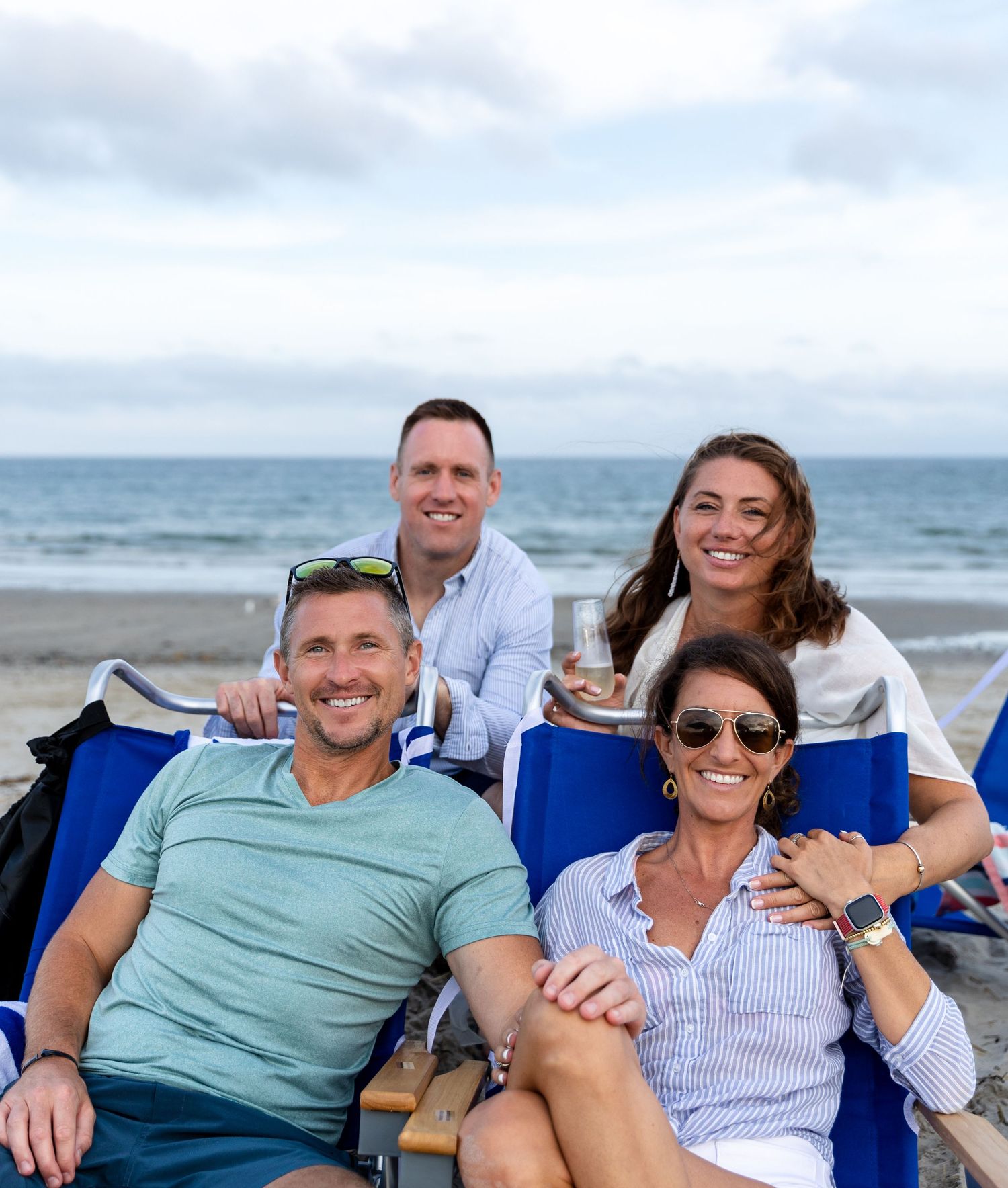 Two couples enjoying a casual beach day together while relaxing in blue chairs by the ocean.