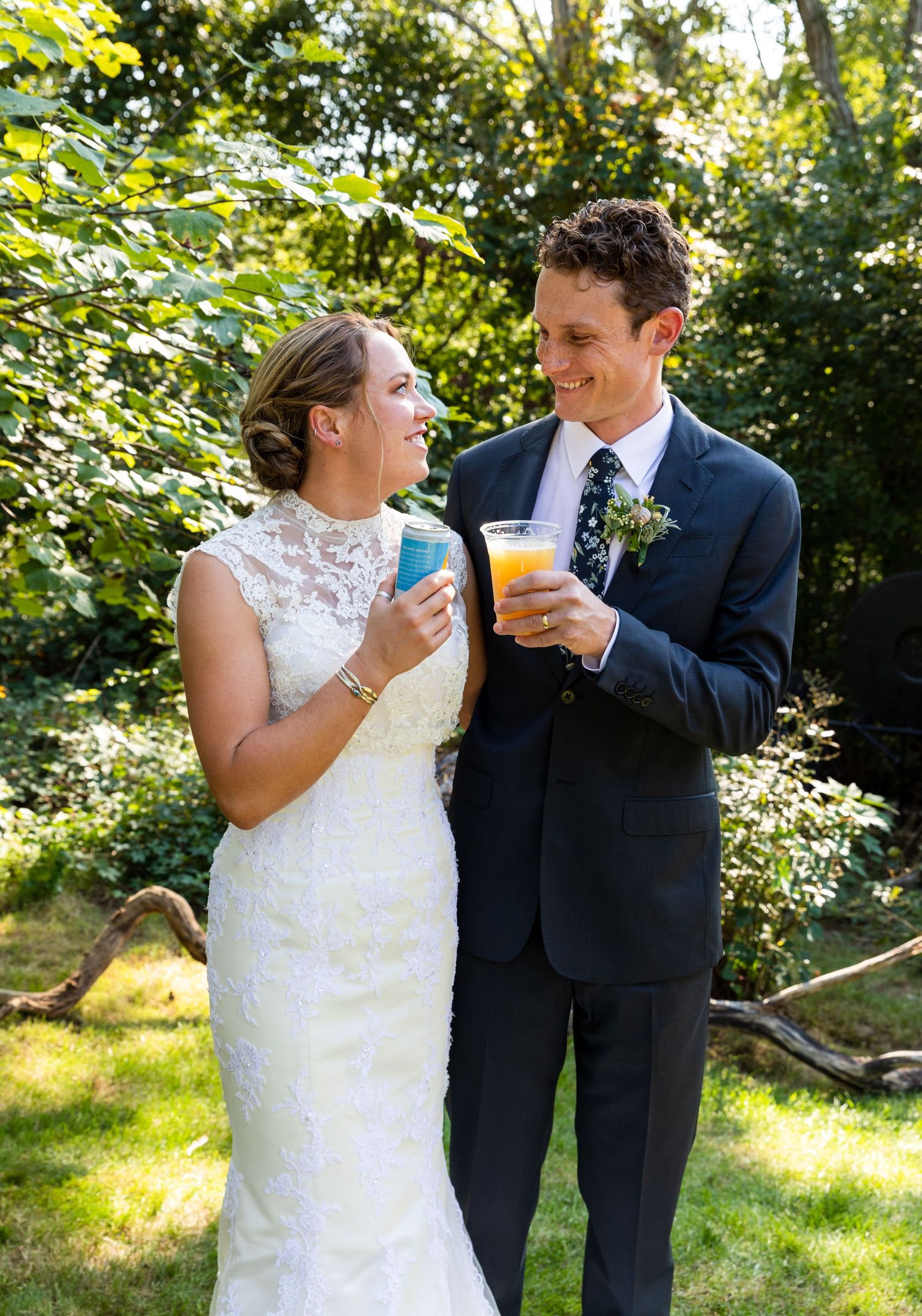 A bride in lace and groom in navy share colorful drinks during an outdoor garden wedding celebration.