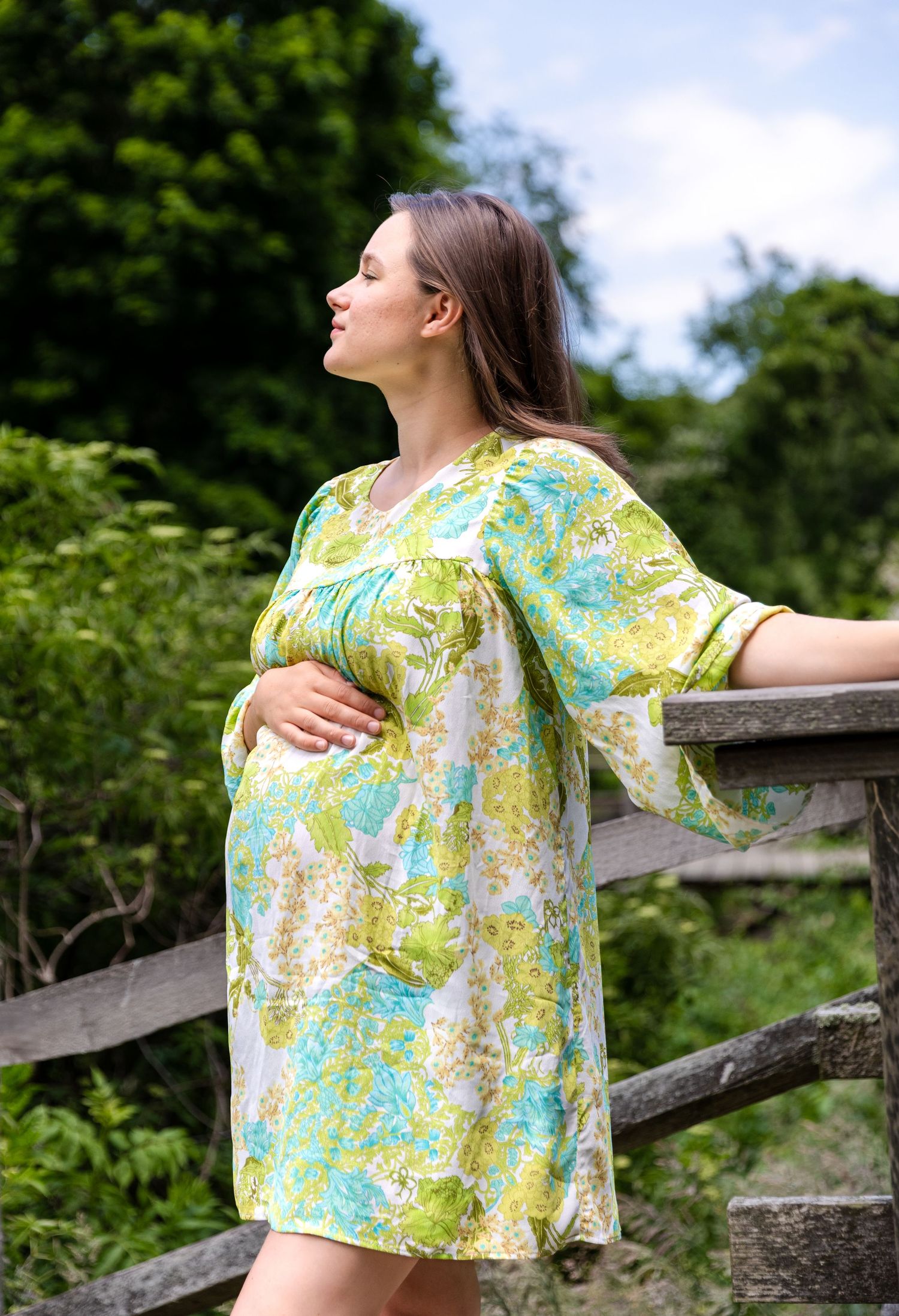 A pregnant woman in a floral print yellow and turquoise dress stands beside a wooden fence on a summer day.