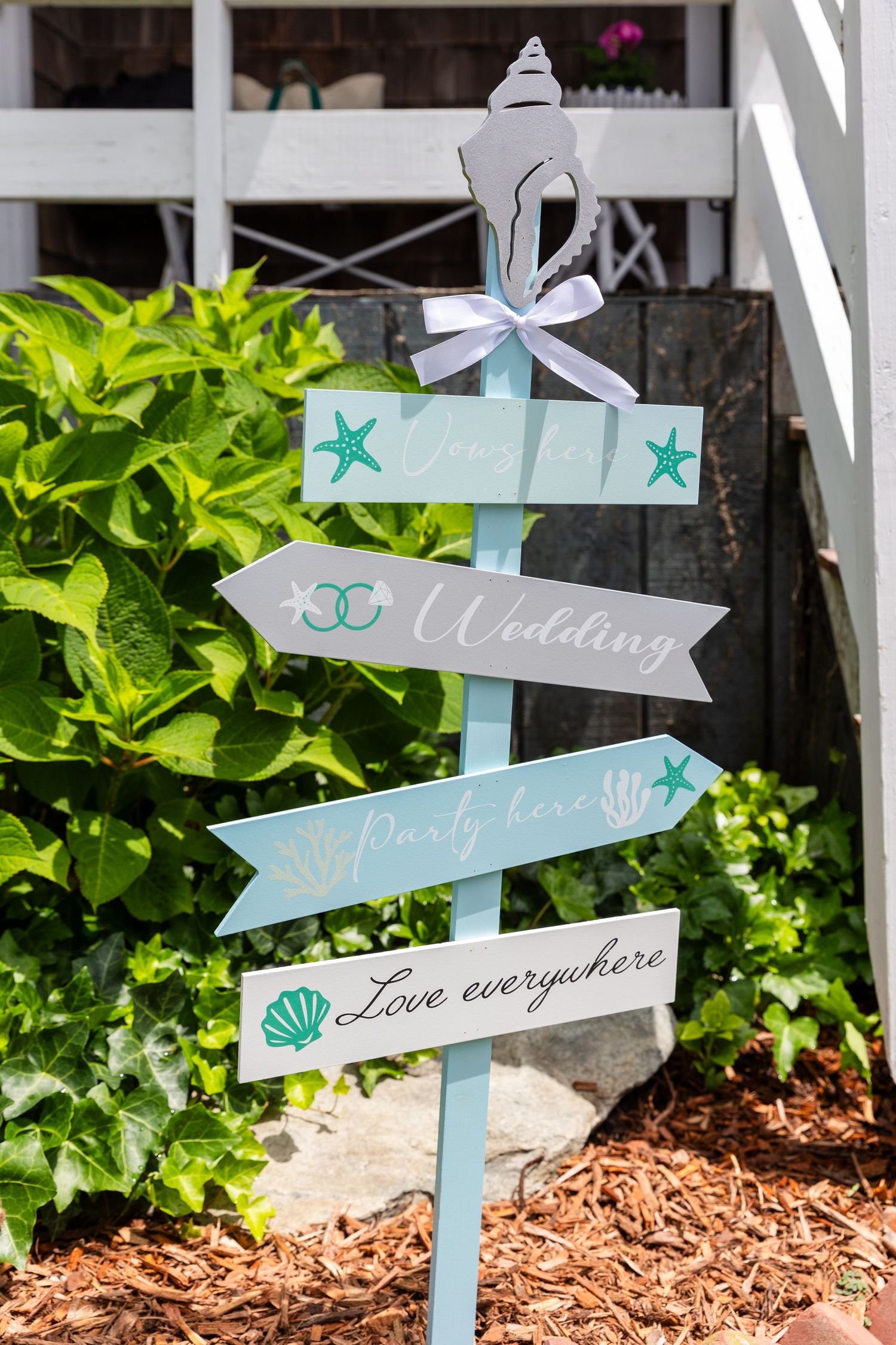 Light blue directional signs with seashell designs for a beach themed wedding displayed against lush green plants and mulch.