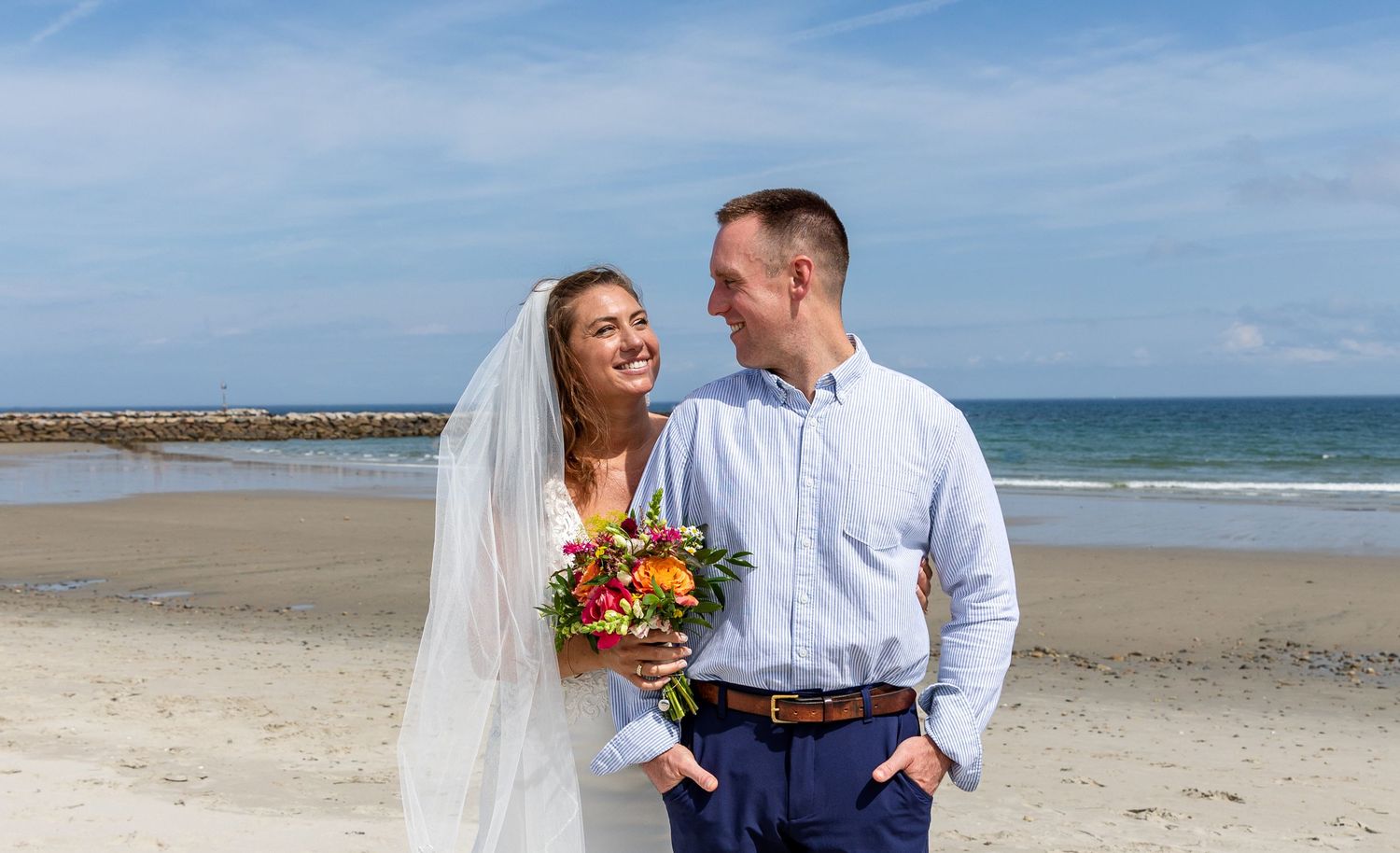 A newlywed couple shares an intimate moment while walking along a scenic beach with blue skies and ocean waves.