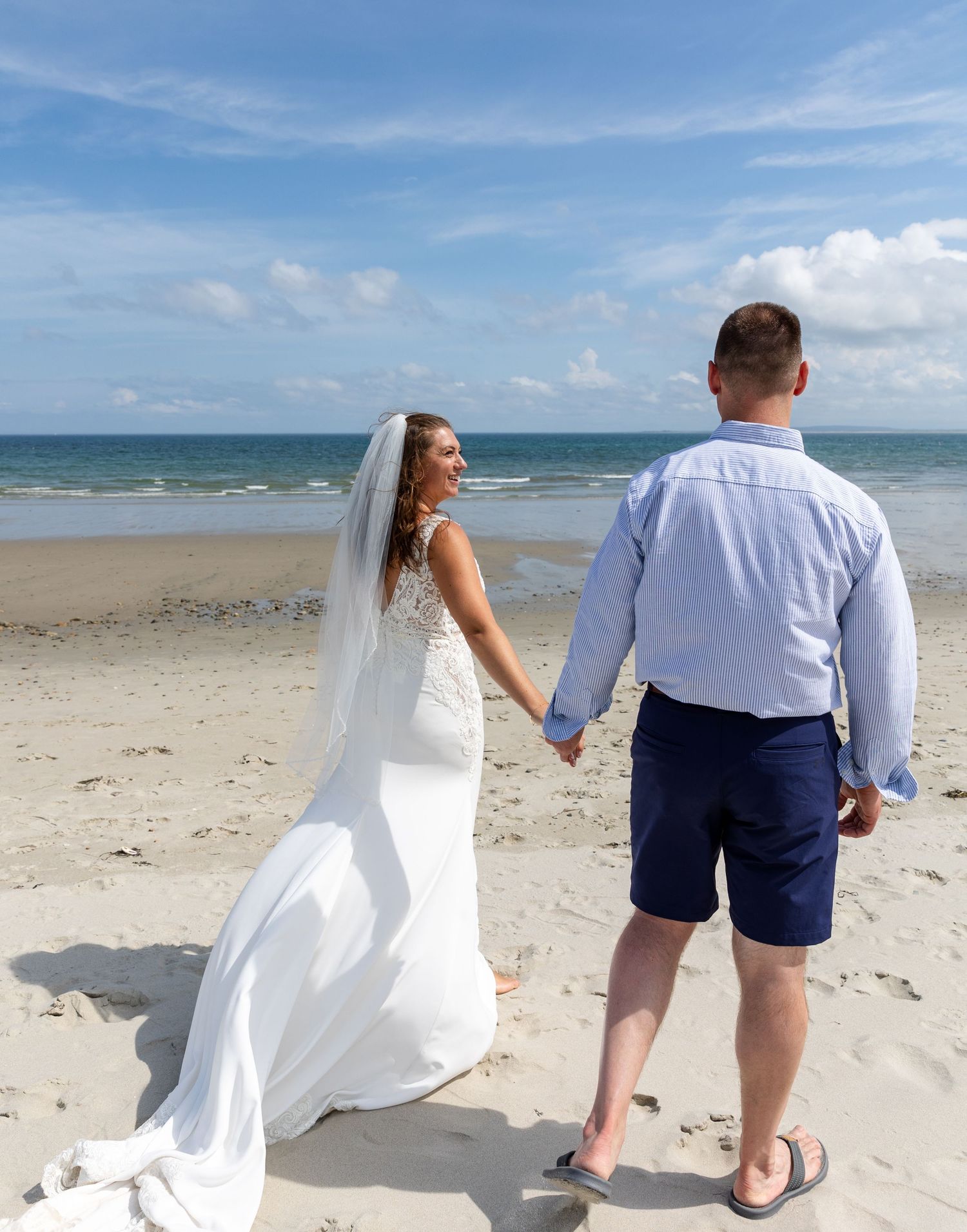 A newly married couple strolls together on a scenic beach with their backs to the camera.