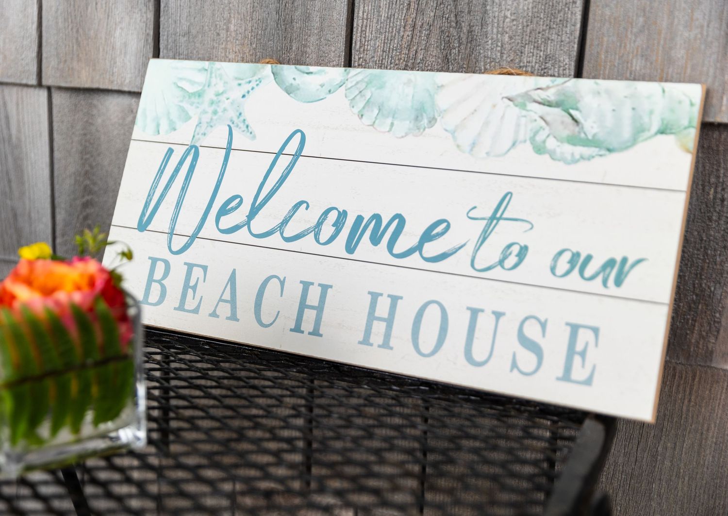 A rustic wooden sign reading 'Welcome to our BEACH HOUSE' with aqua blue accents sits on a patio table near flowers.