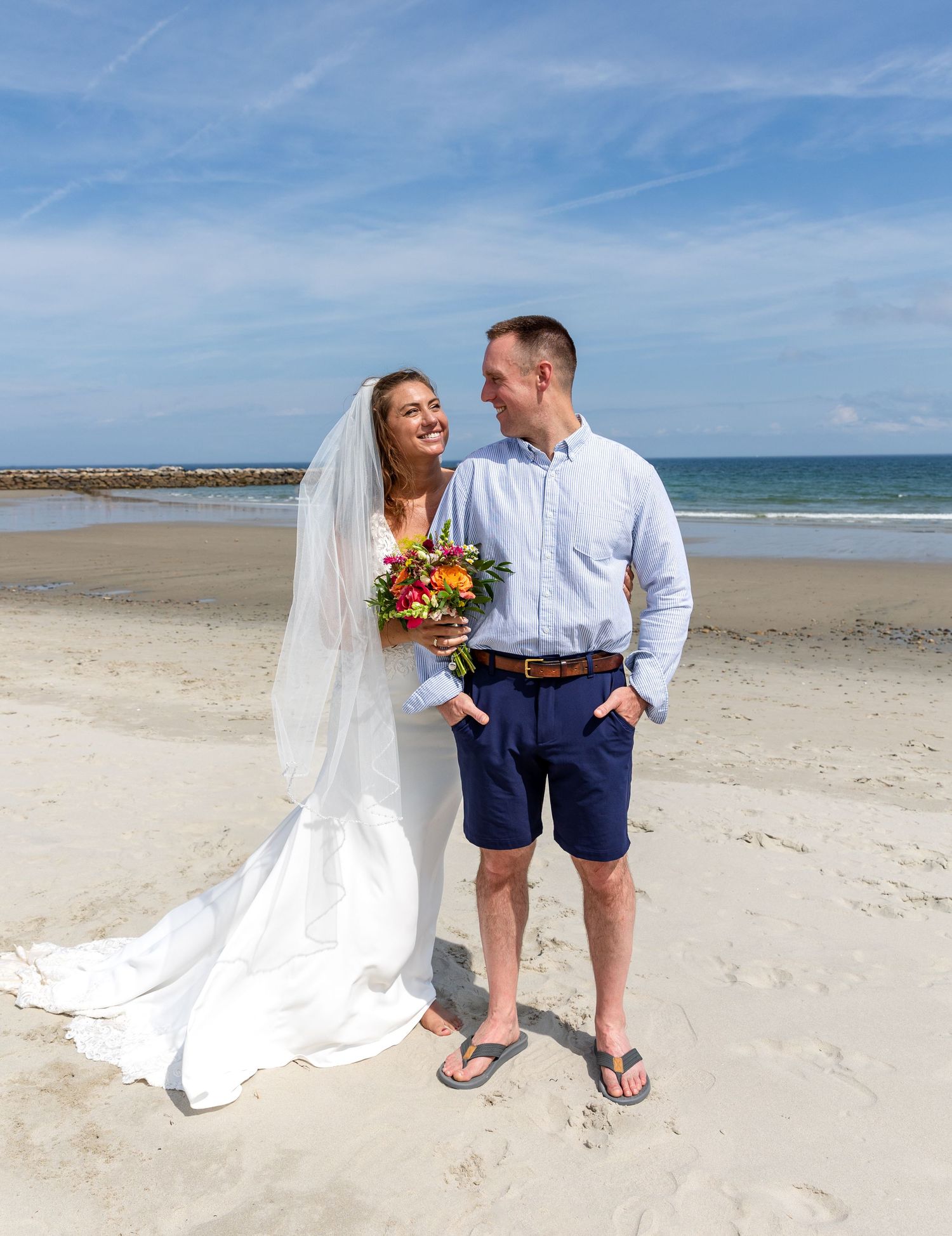 A newly married couple stands together on a sunny beach, with ocean waves and rocks visible in the background.