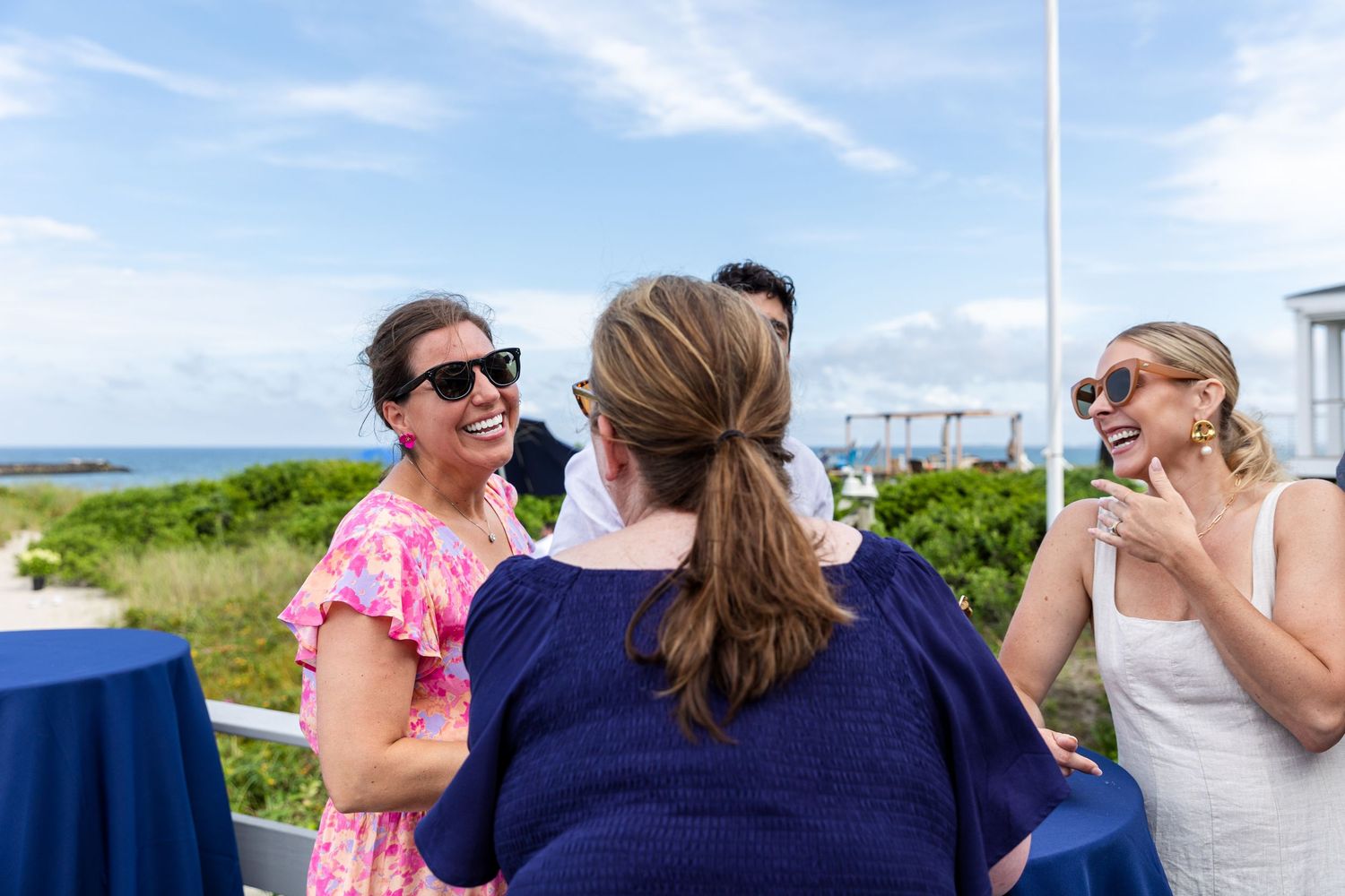 A group of people share a conversation at an outdoor event with blue tablecloths and ocean views in the background.