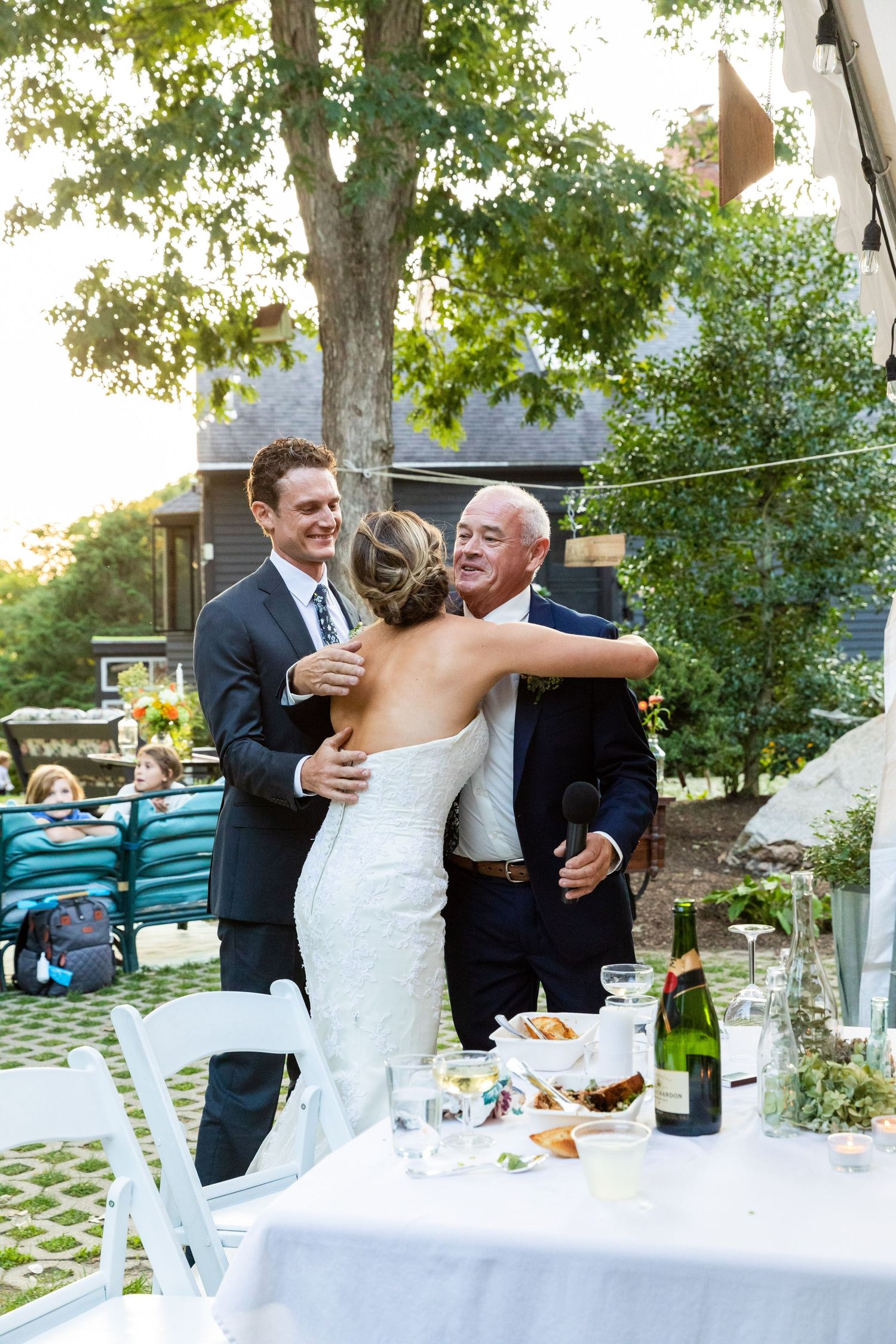 A wedding reception celebration outdoors with a white-clothed table, champagne bottle, and guests embracing by a large tree.
