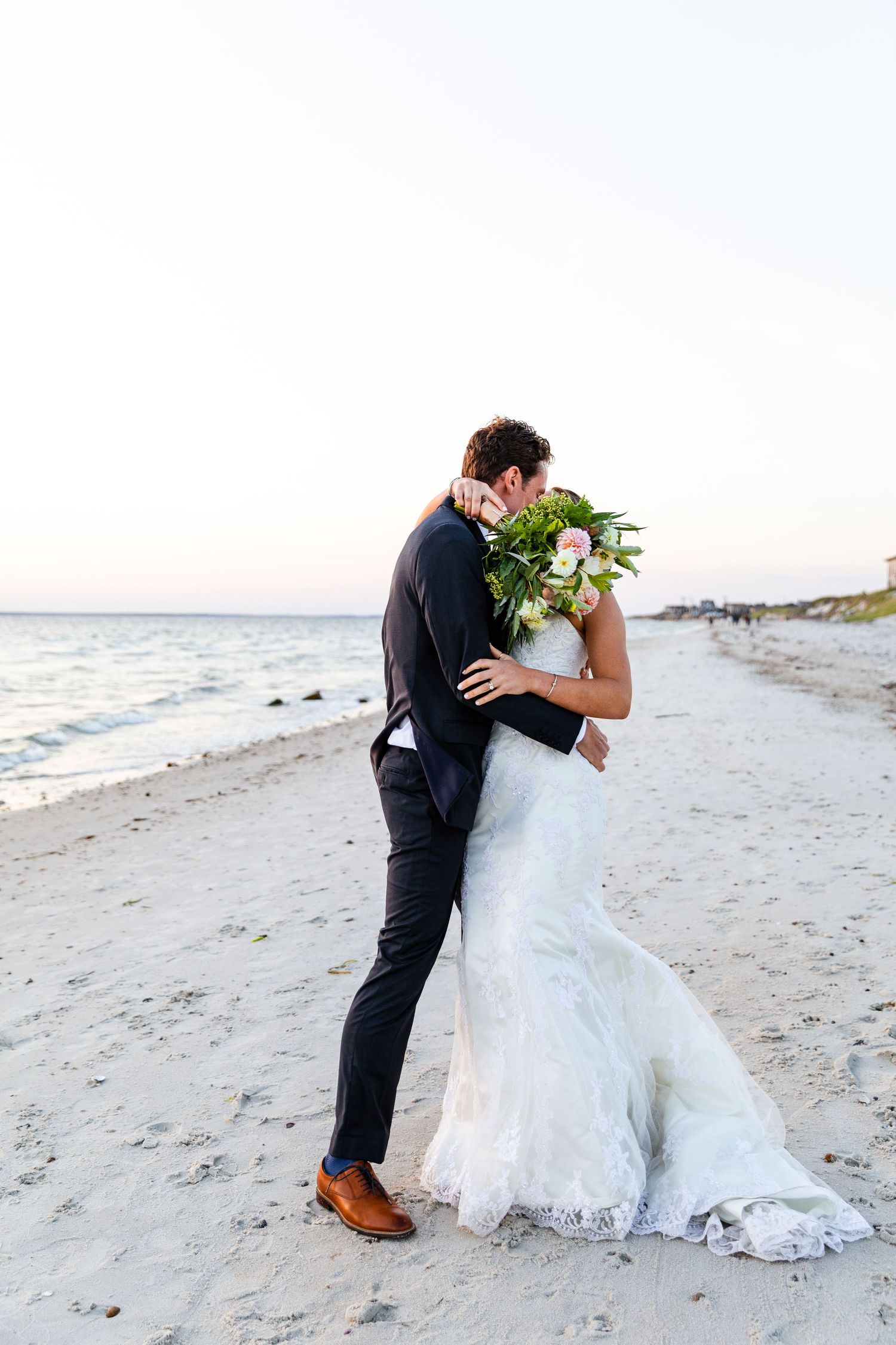 A couple in wedding attire embraces on a sandy beach at sunset with waves and shoreline in the background.