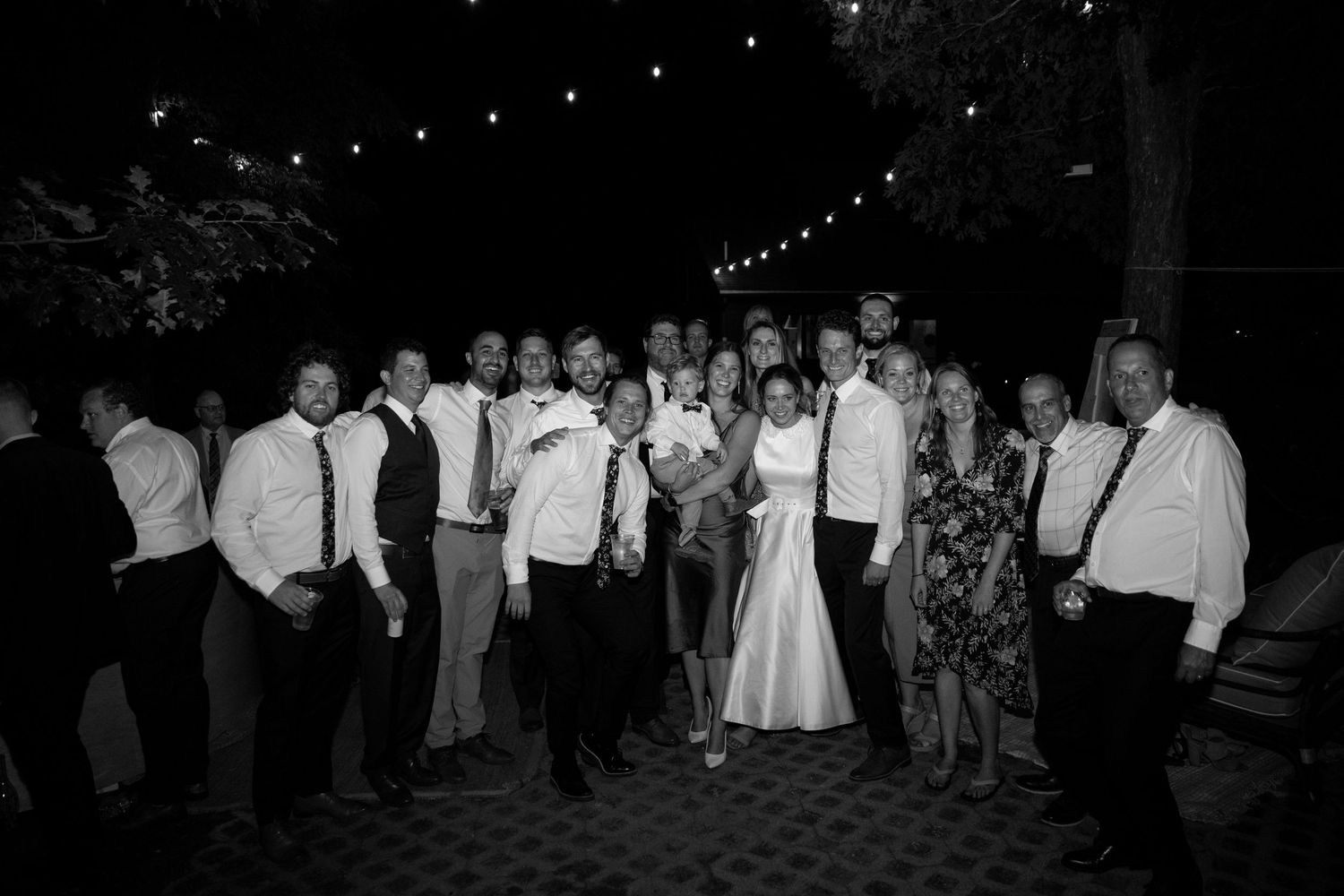 A black and white photo shows wedding guests dancing in a circle under string lights at an outdoor reception.