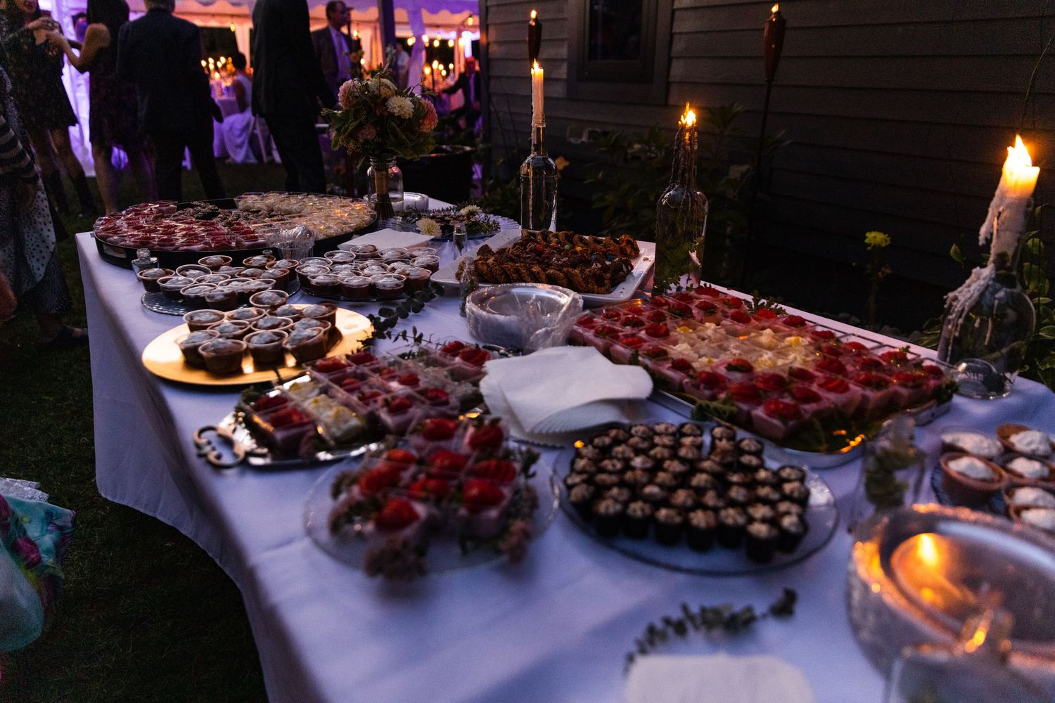 An elegant dessert buffet table features assorted cakes, chocolates and treats displayed on white linens with soft lighting.