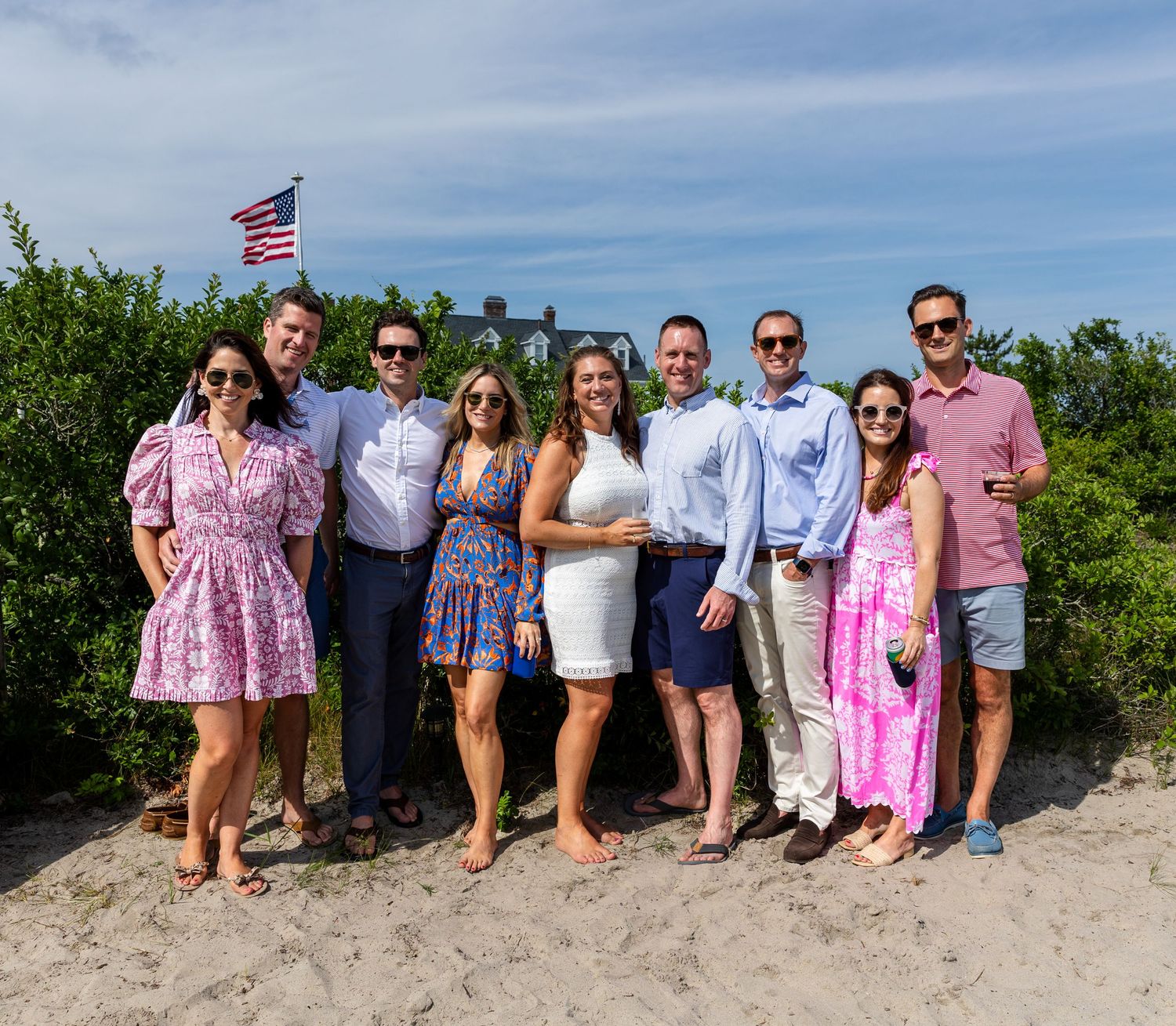 A group in summer attire poses together on a sandy beach with an American flag waving in the background.