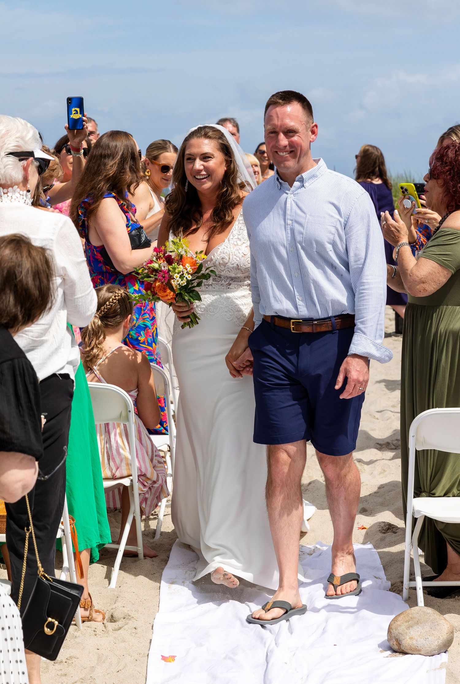 A beach wedding celebration with guests and attendees gathered on the sand on a sunny day with blue skies.