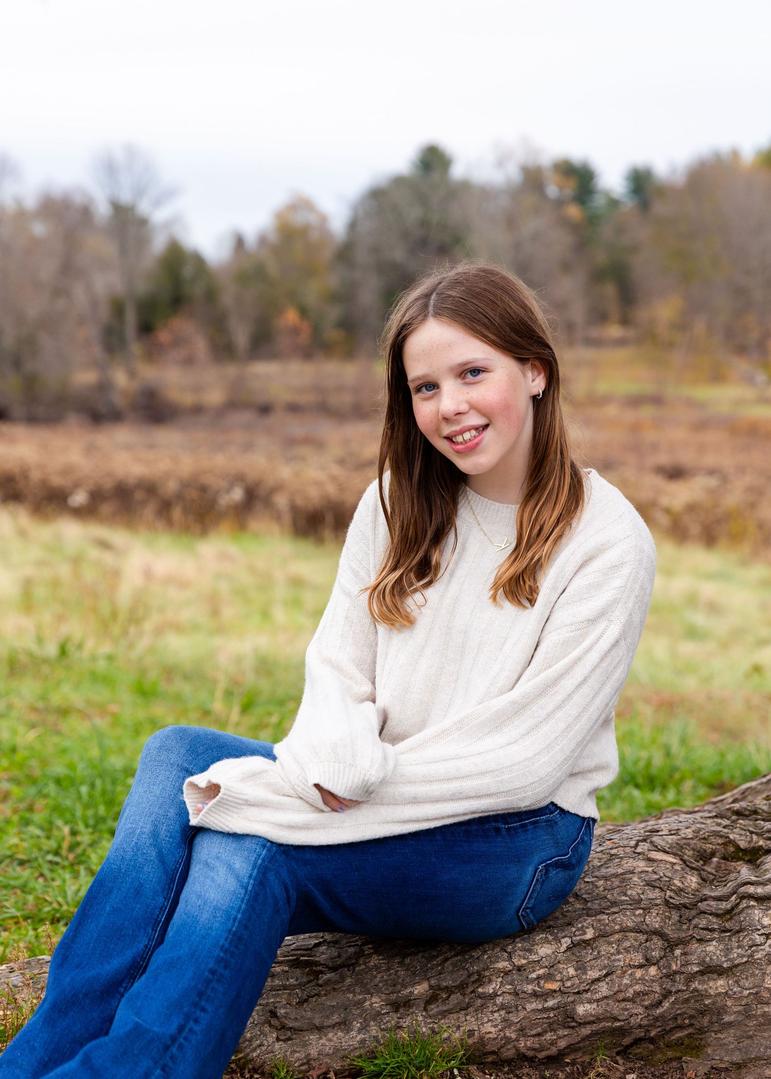 A person in a cream sweater and jeans sits casually on a fallen log in an autumn field with trees in the distance.