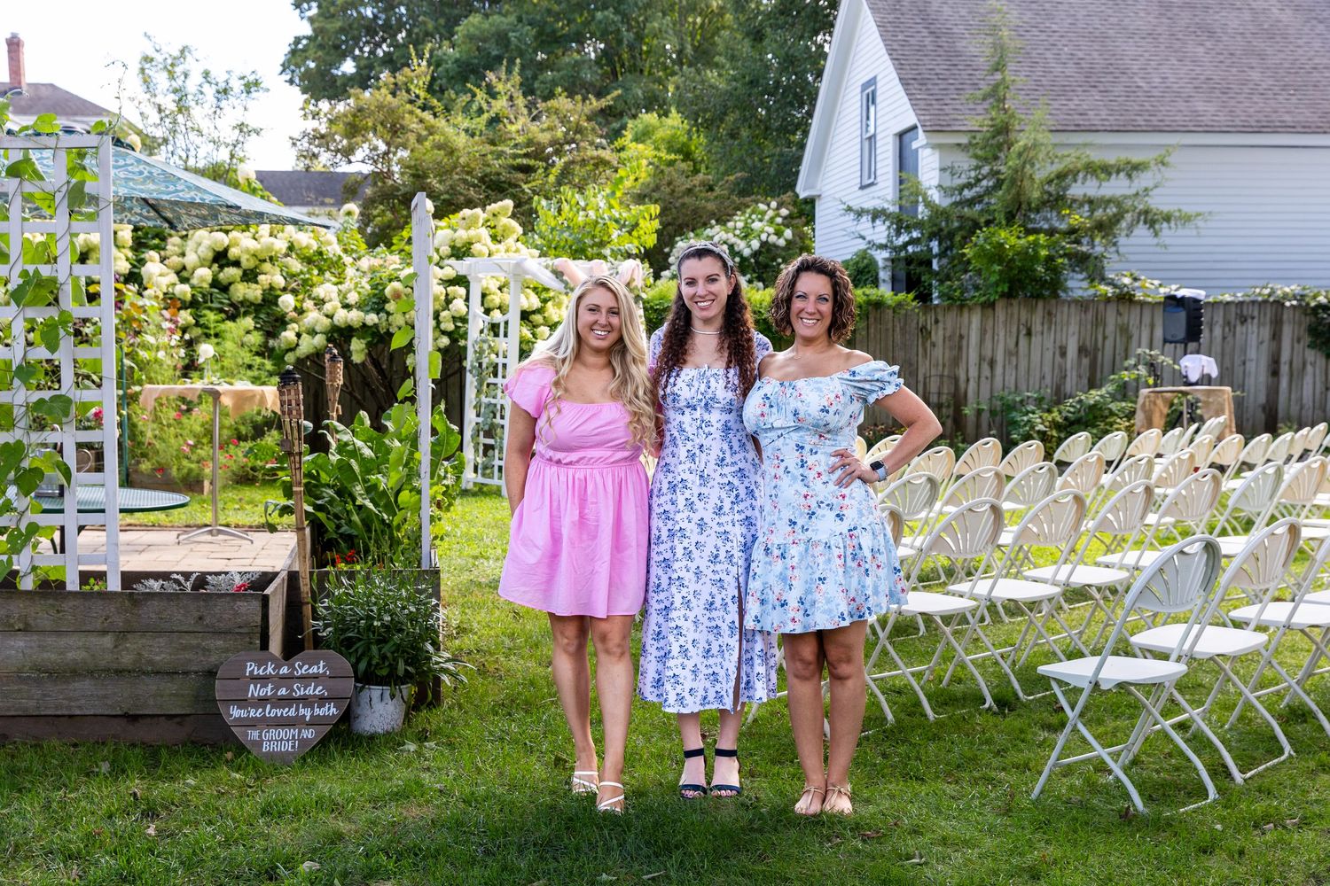 Three people stand in front of a white garden arbor with chairs set up on a lawn for an outdoor event.