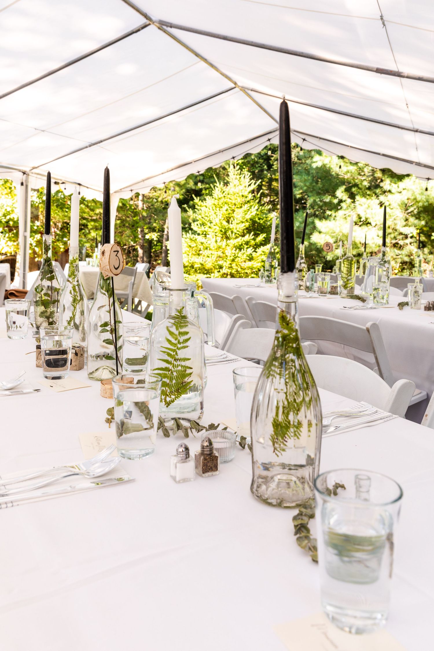 Elegant white event tent interior with glass vases and ferns arranged on minimalist tables for a wedding reception setup.