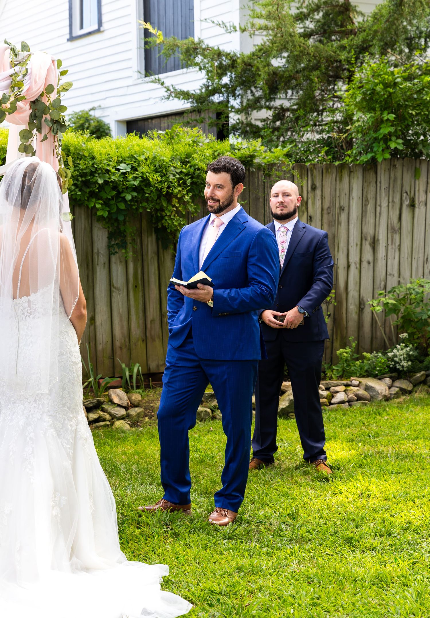 Wedding party in navy blue suits gathers outdoors on grass with a white fence and greenery in the background.