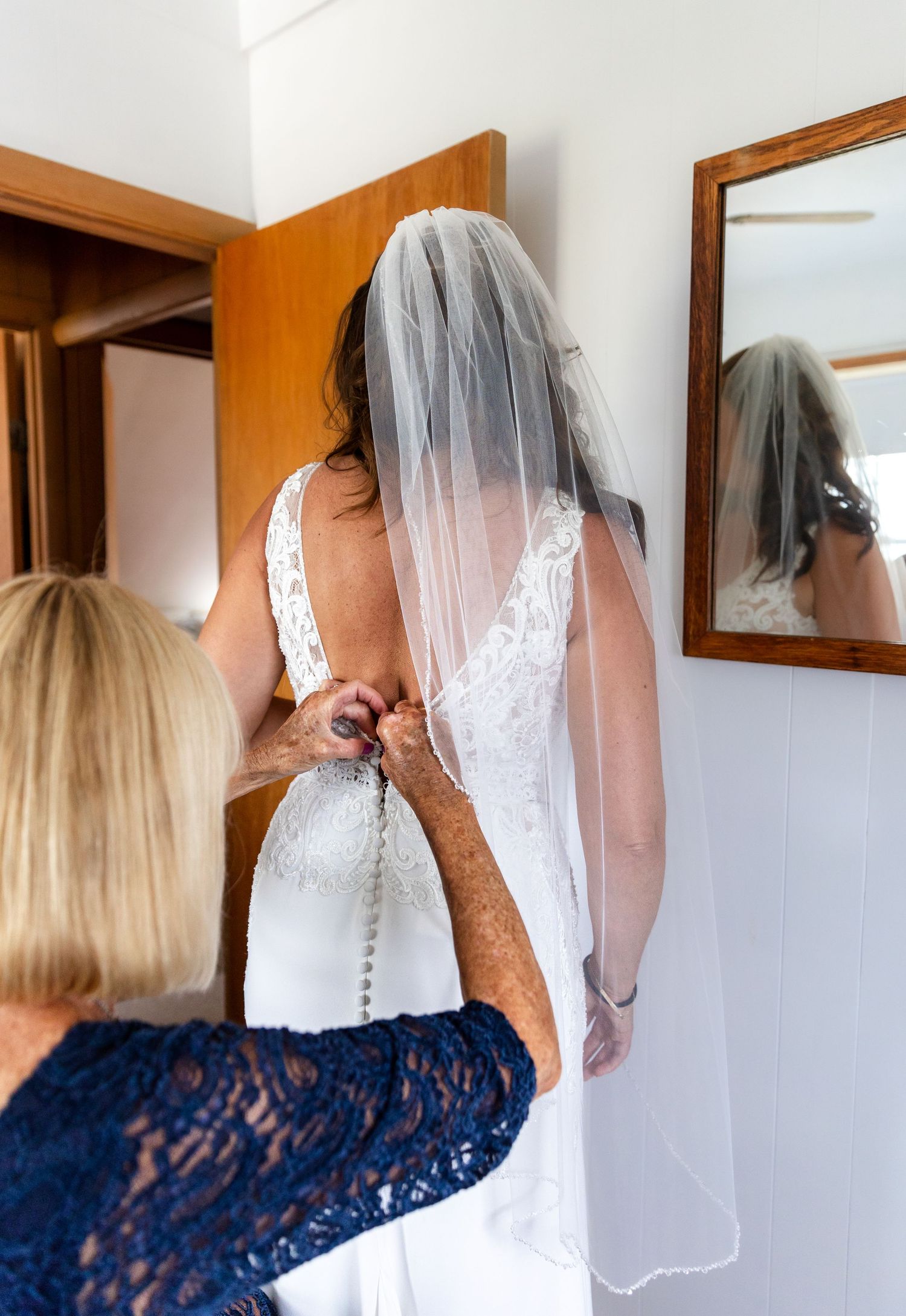 Someone in a white wedding dress with veil gets help with buttons from a person wearing a navy blue lace dress.