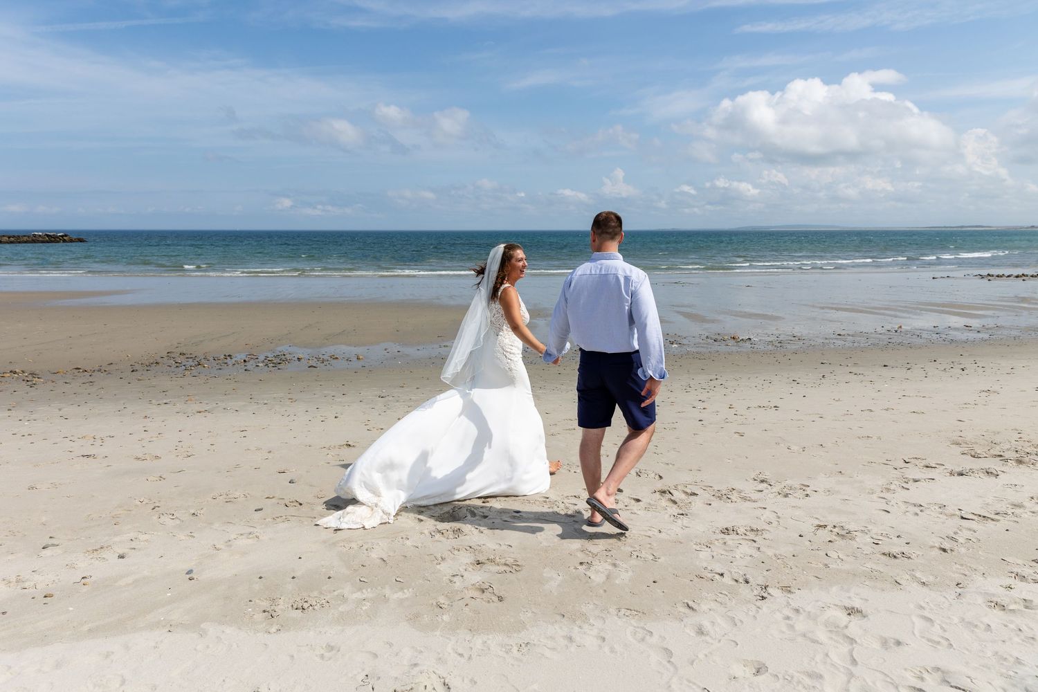 A couple walks together on a scenic white sandy beach with gentle ocean waves and blue skies in the background.