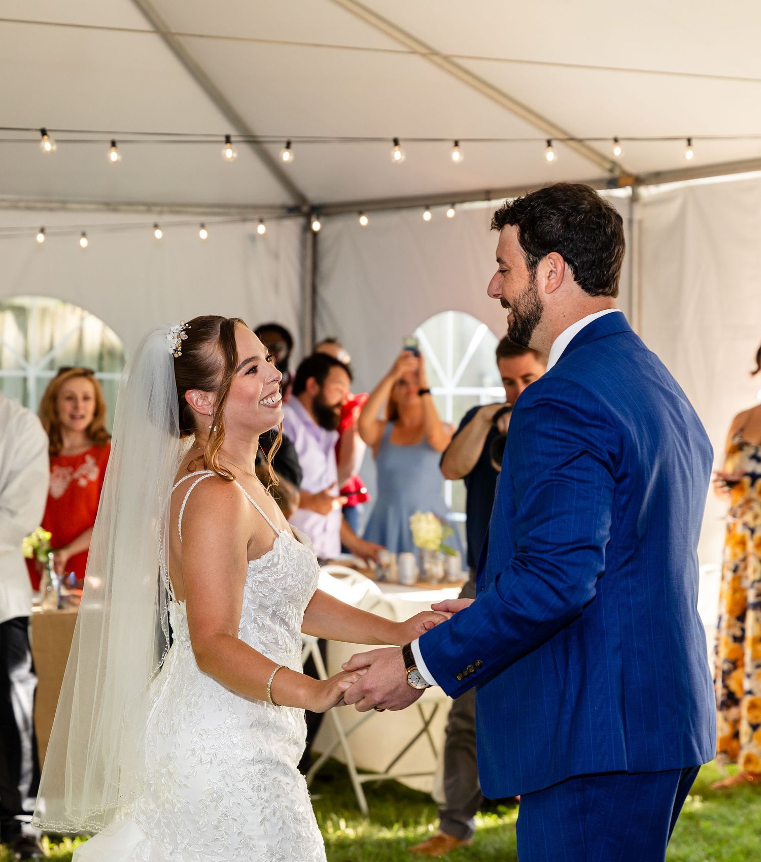 A couple shares a joyful moment during their first dance at an outdoor tented wedding reception with string lights overhead.