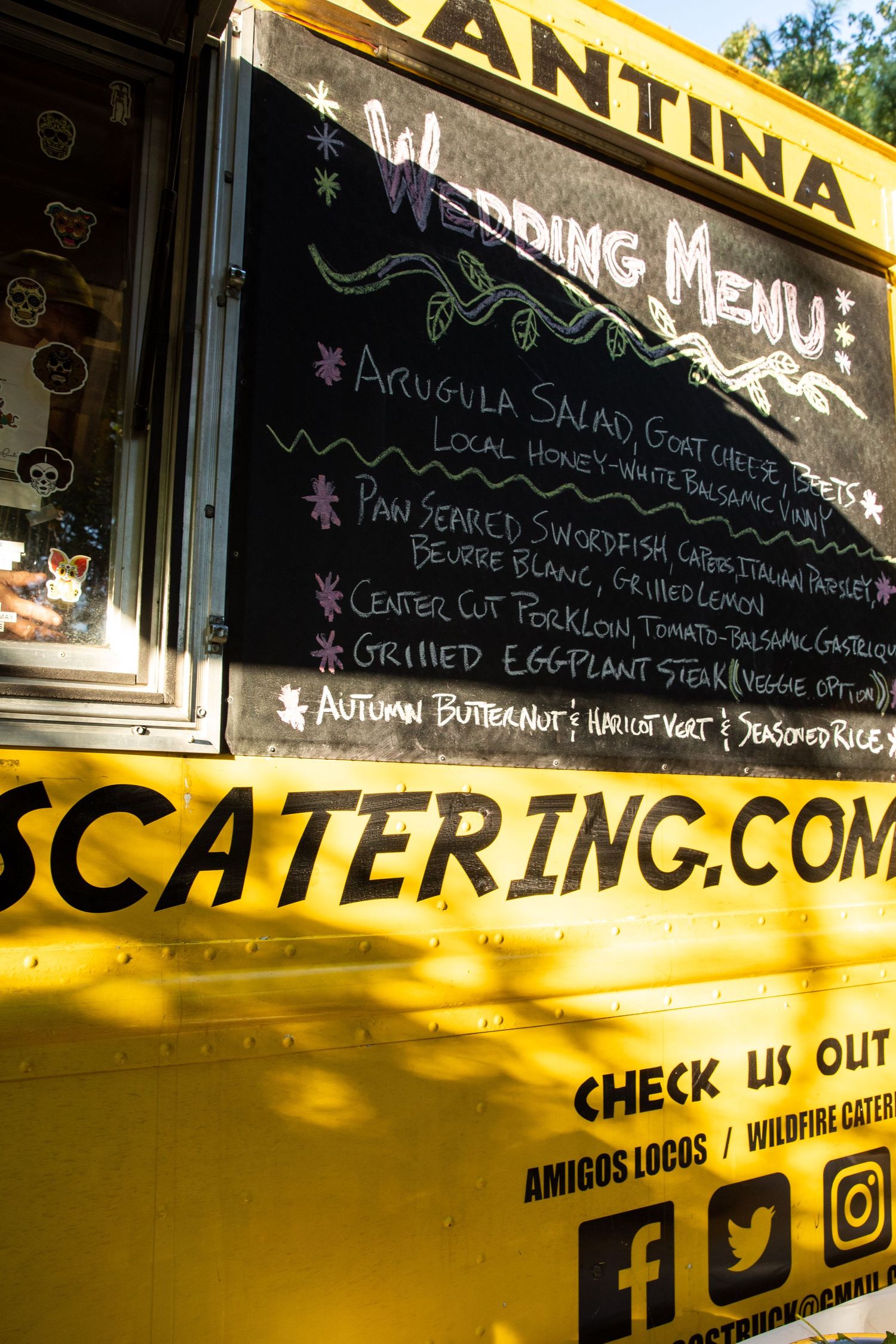 Yellow food truck with wedding menu items written on chalkboard sign and social media links displayed below.