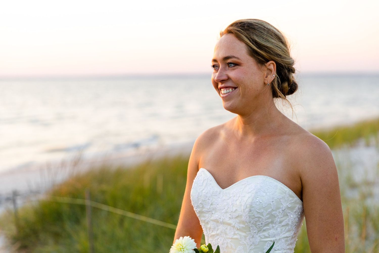 A bride in a strapless white wedding dress smiles while standing by the beach at sunset.