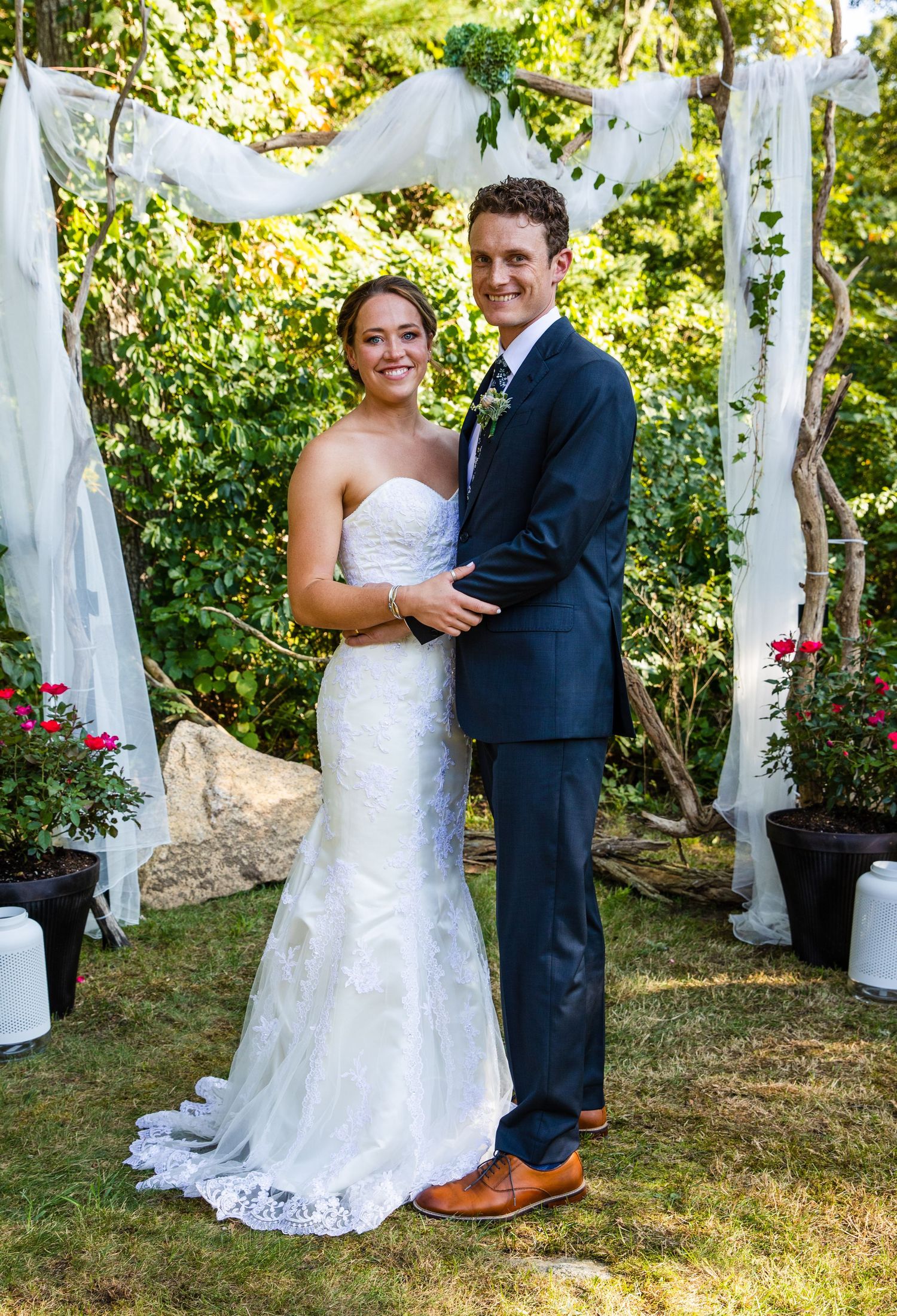 Newlywed couple poses beneath flowing white fabric and floral decorations at outdoor garden wedding ceremony.
