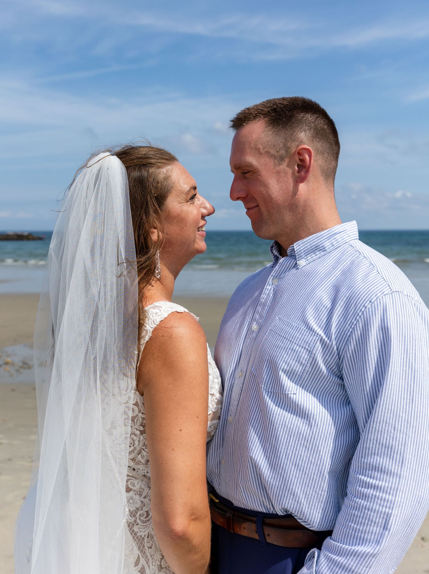 A couple stands together on a sunny beach, sharing joyful laughter while wearing wedding attire near the ocean.