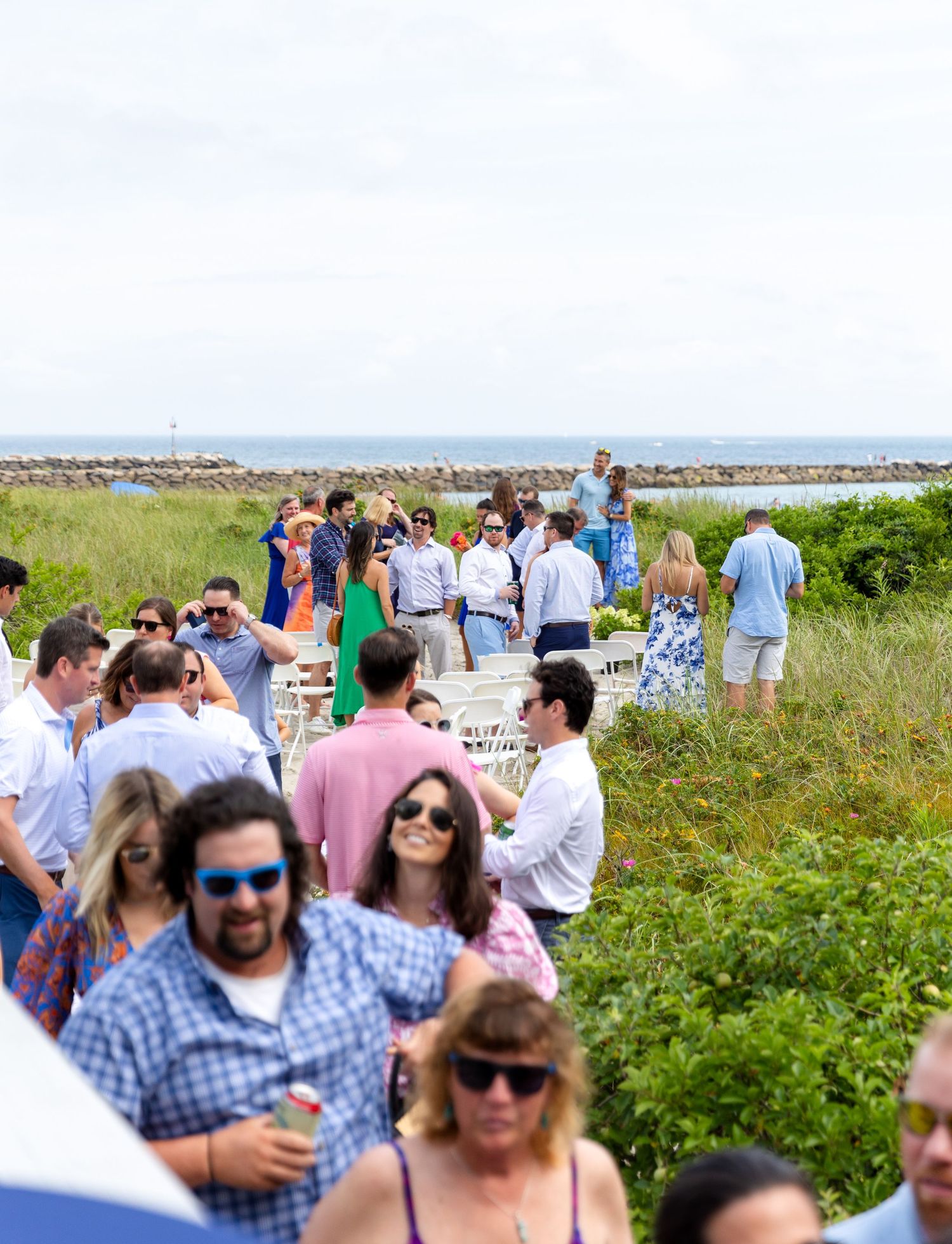 A casual outdoor gathering of people mingling on a grassy path near the ocean with a stone jetty in the background.